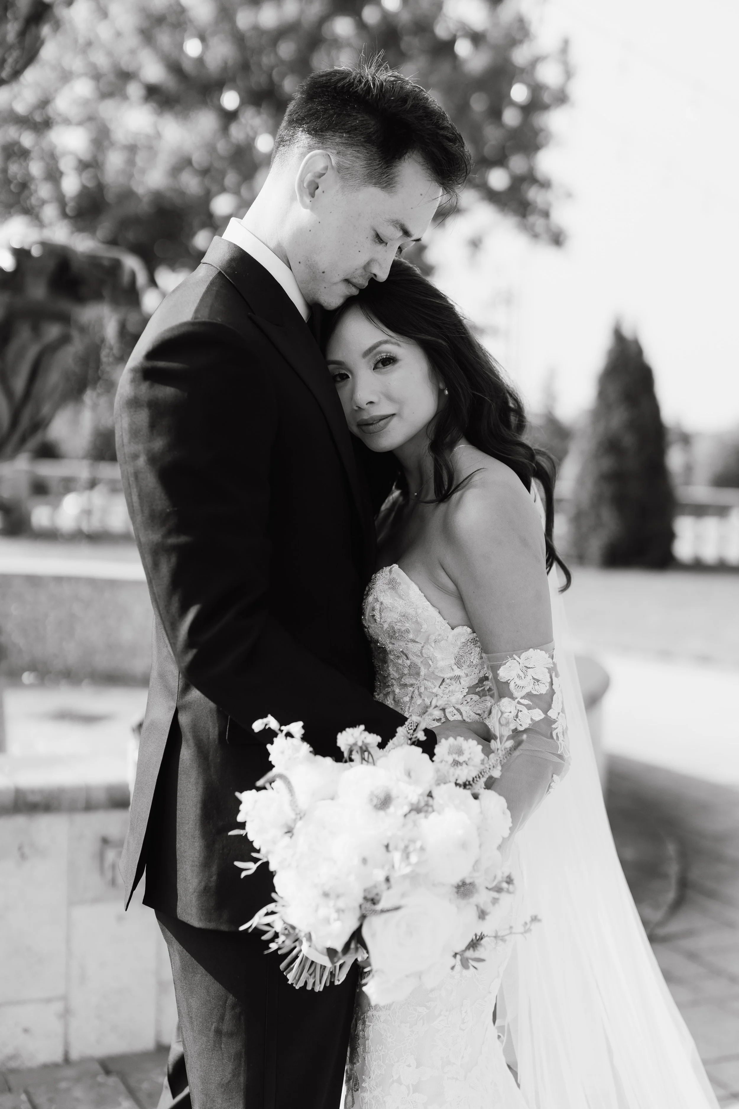 Black and white photo of a bride and groom embracing outdoors. The groom has short dark hair and is wearing a dark suit, while the bride has long dark hair and is wearing a lace wedding dress. The bride holds a bouquet of flowers and gazes at the cam