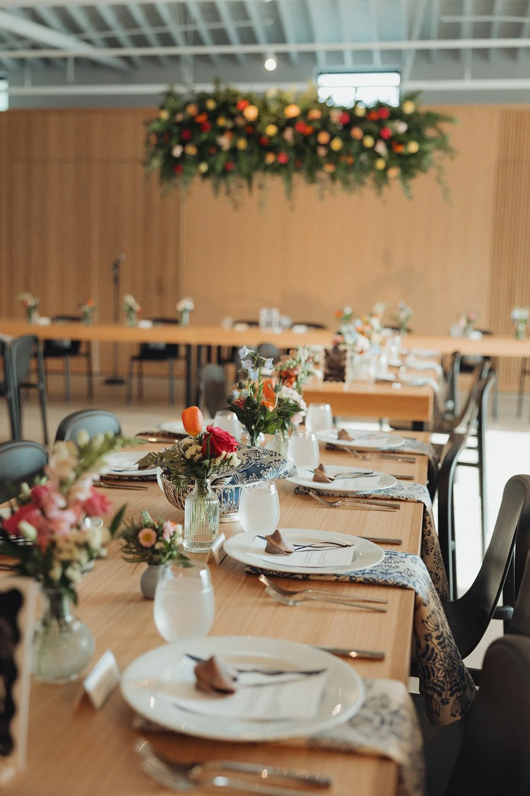 A long dining table set with plates, silverware, folded napkins, and water glasses, decorated with colorful floral centerpieces in vases, in a spacious room with wooden walls and a floral arrangement hanging from the ceiling.