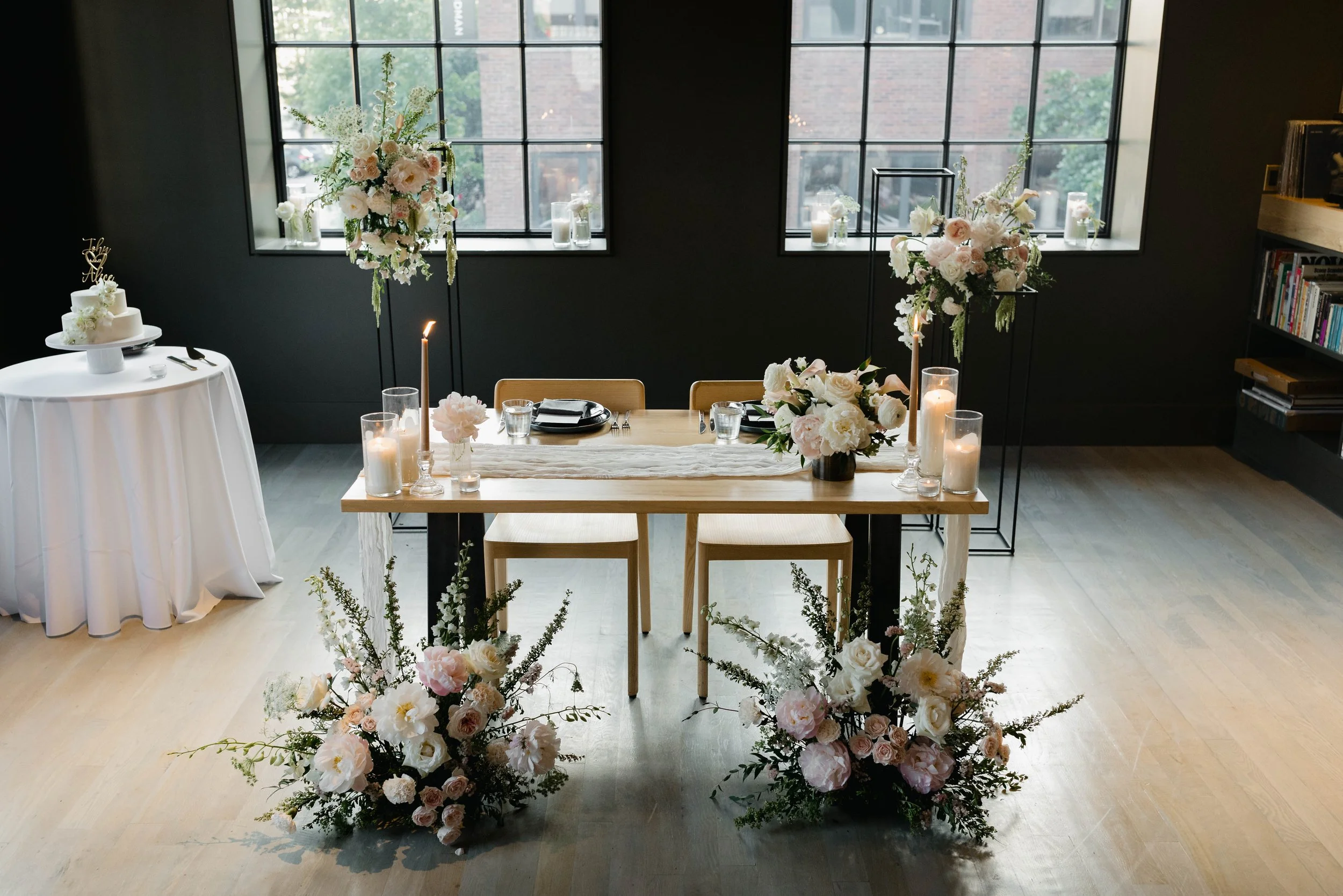 Elegant wedding or event reception setup with floral arrangements, candles, and a decorated table in front of large window panes.