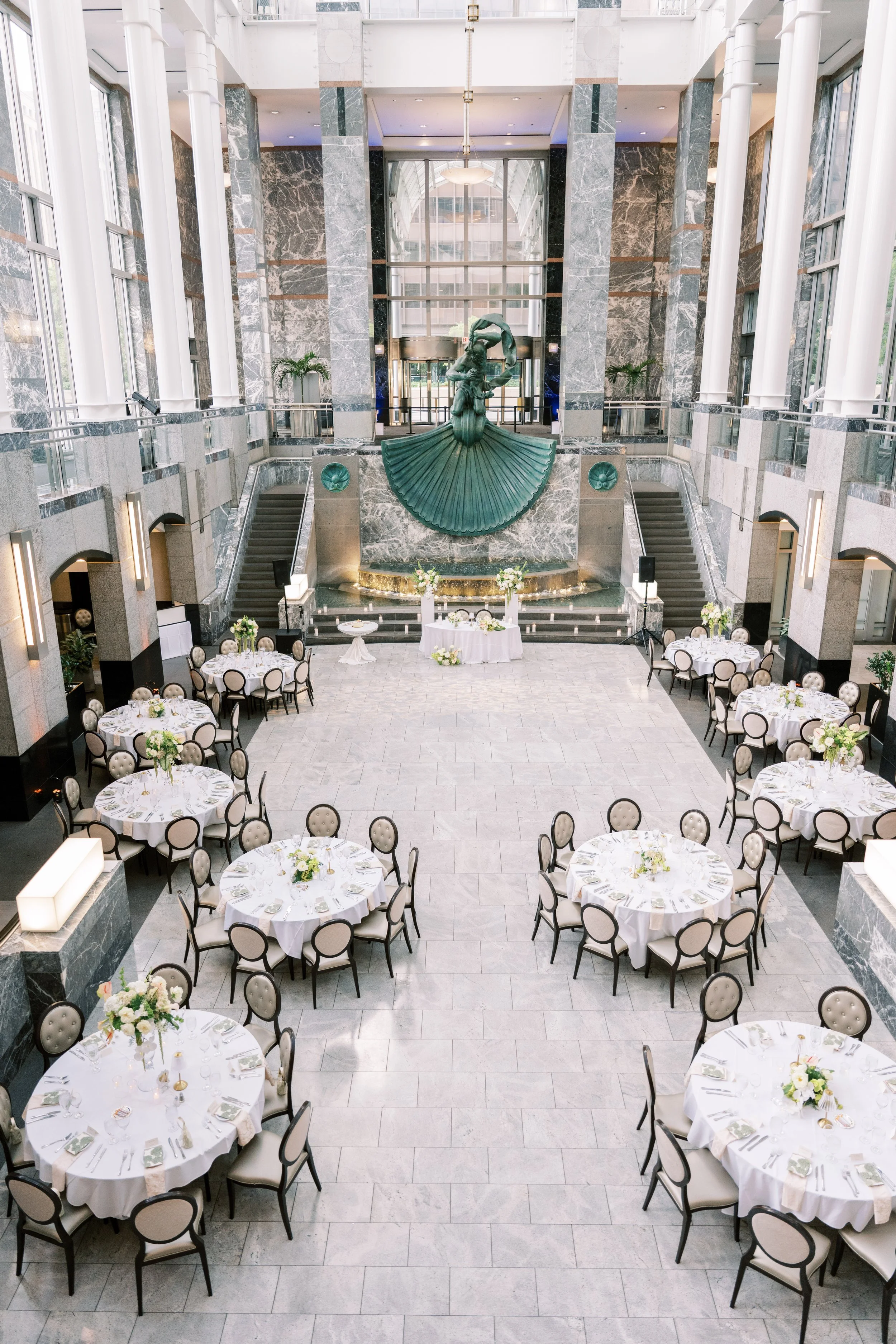 Elegant banquet hall with round tables covered in white tablecloths, decorated with white flowers, set for a formal event. The hall features high ceilings, large windows, and a marble staircase on each side leading to an upper level. A large abstract