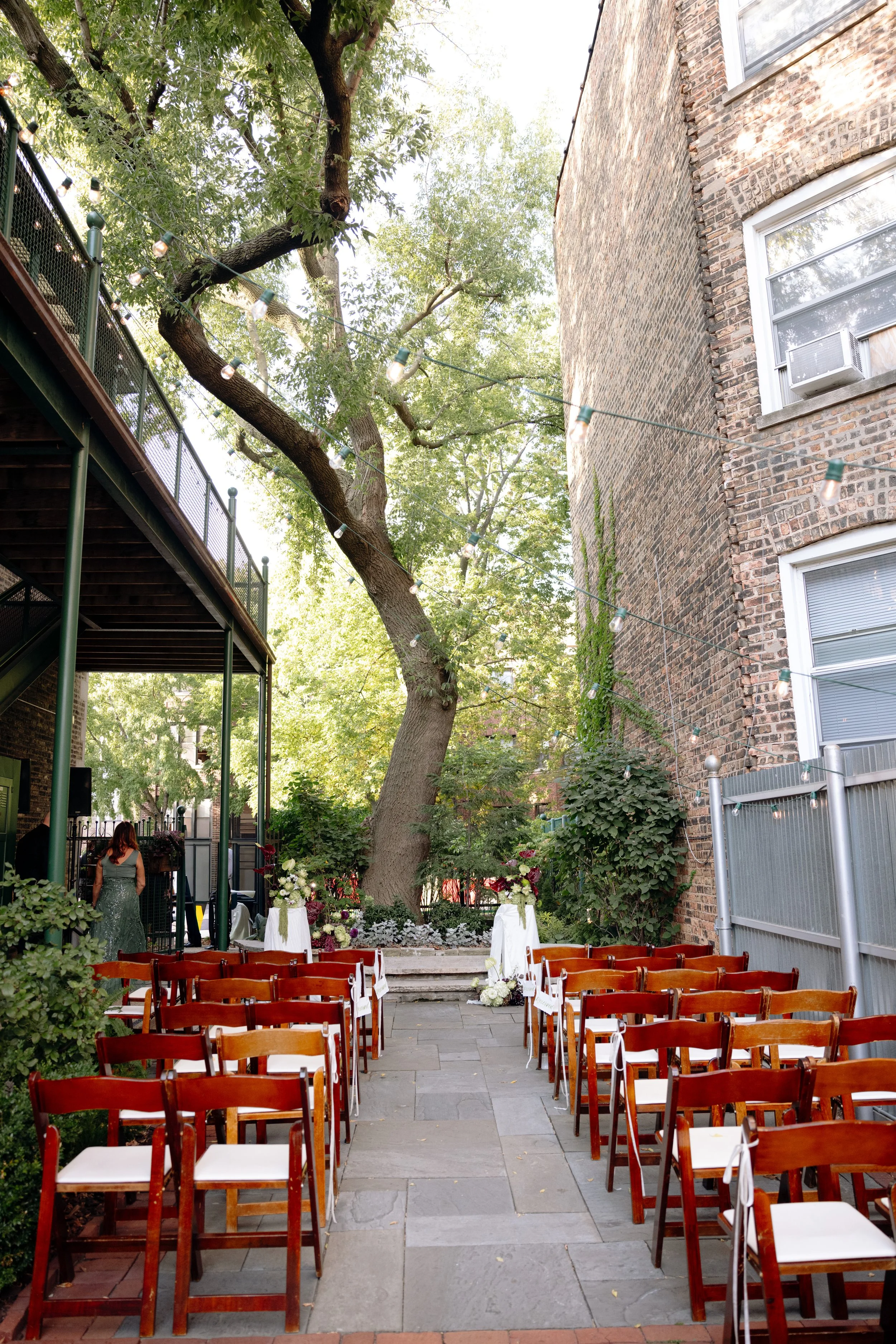 Outdoor wedding setup in a courtyard with rows of wooden chairs, floral arrangements, trees, string lights, and brick buildings.