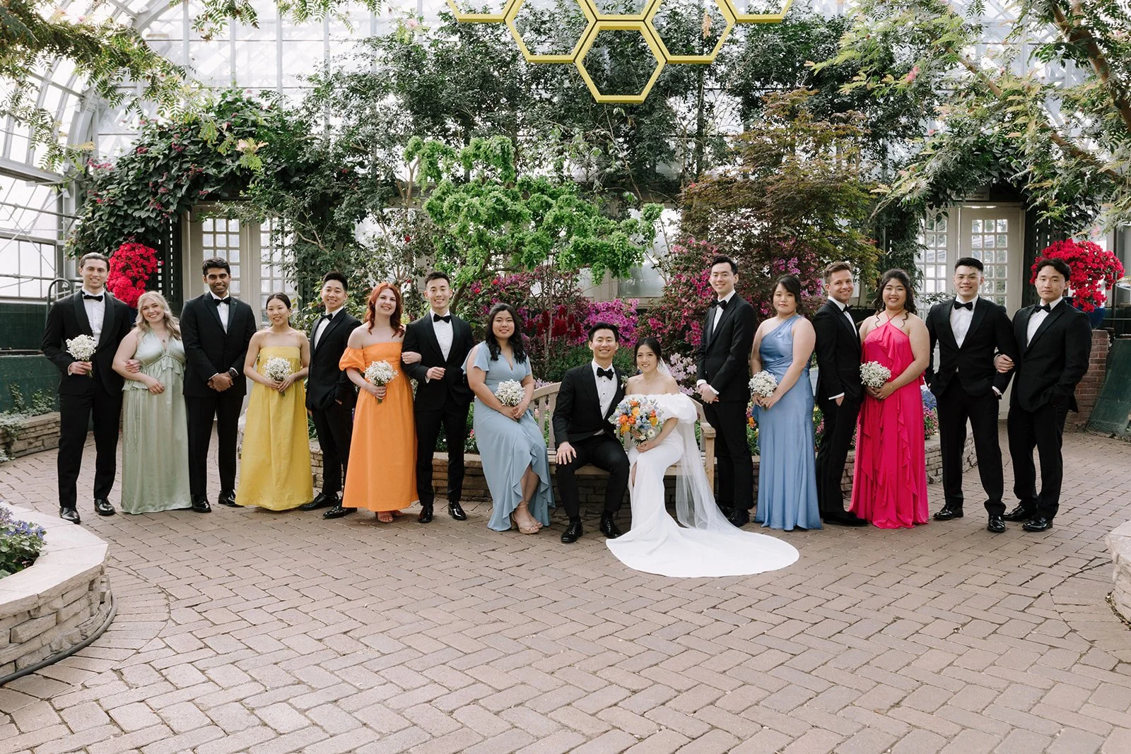 Group of people, including a bride and groom, posing for a photo in a greenhouse with lush greenery and flowers.