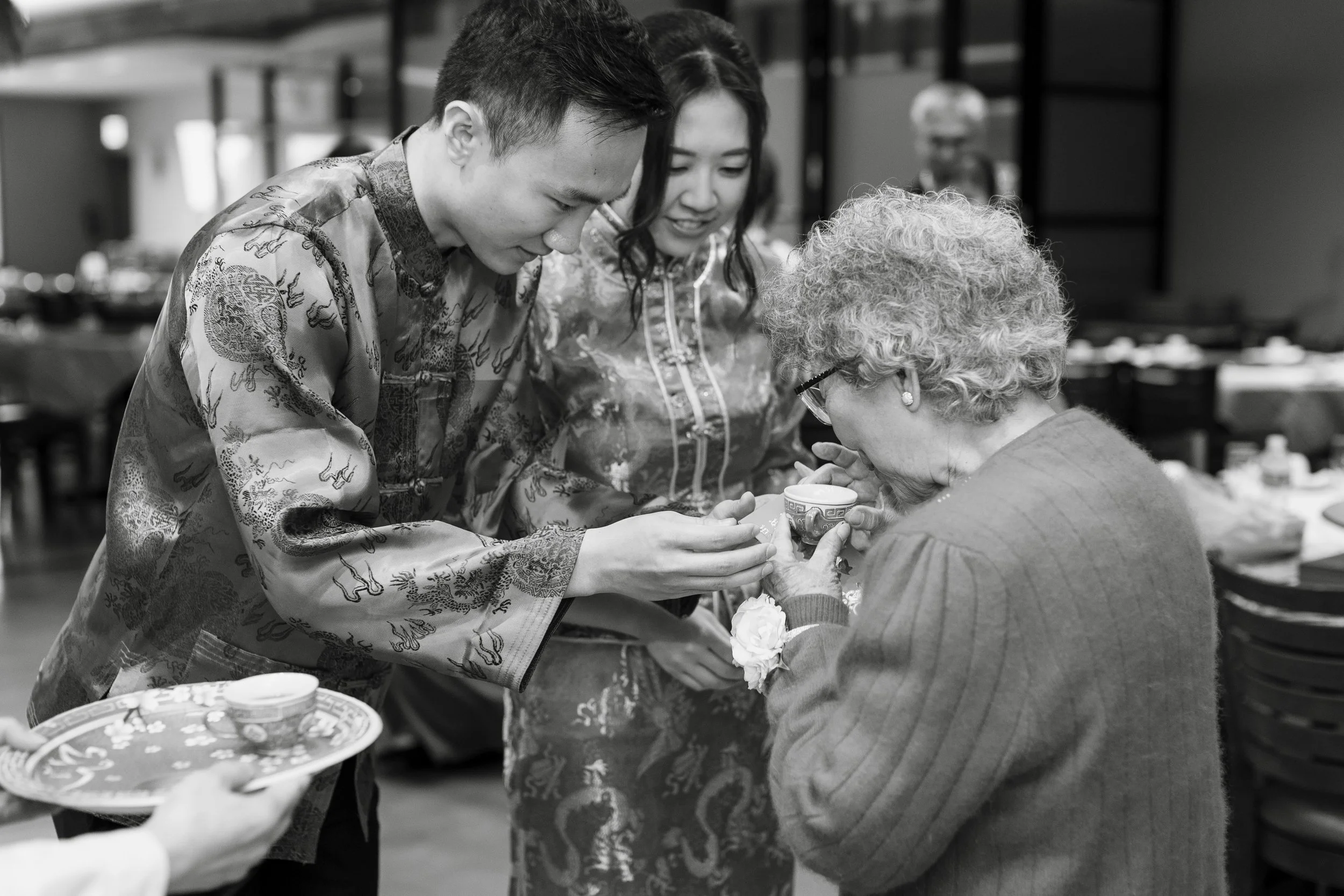 A young man and woman in traditional Chinese attire sharing a cup with an elderly woman, likely in a cultural or family gathering setting.