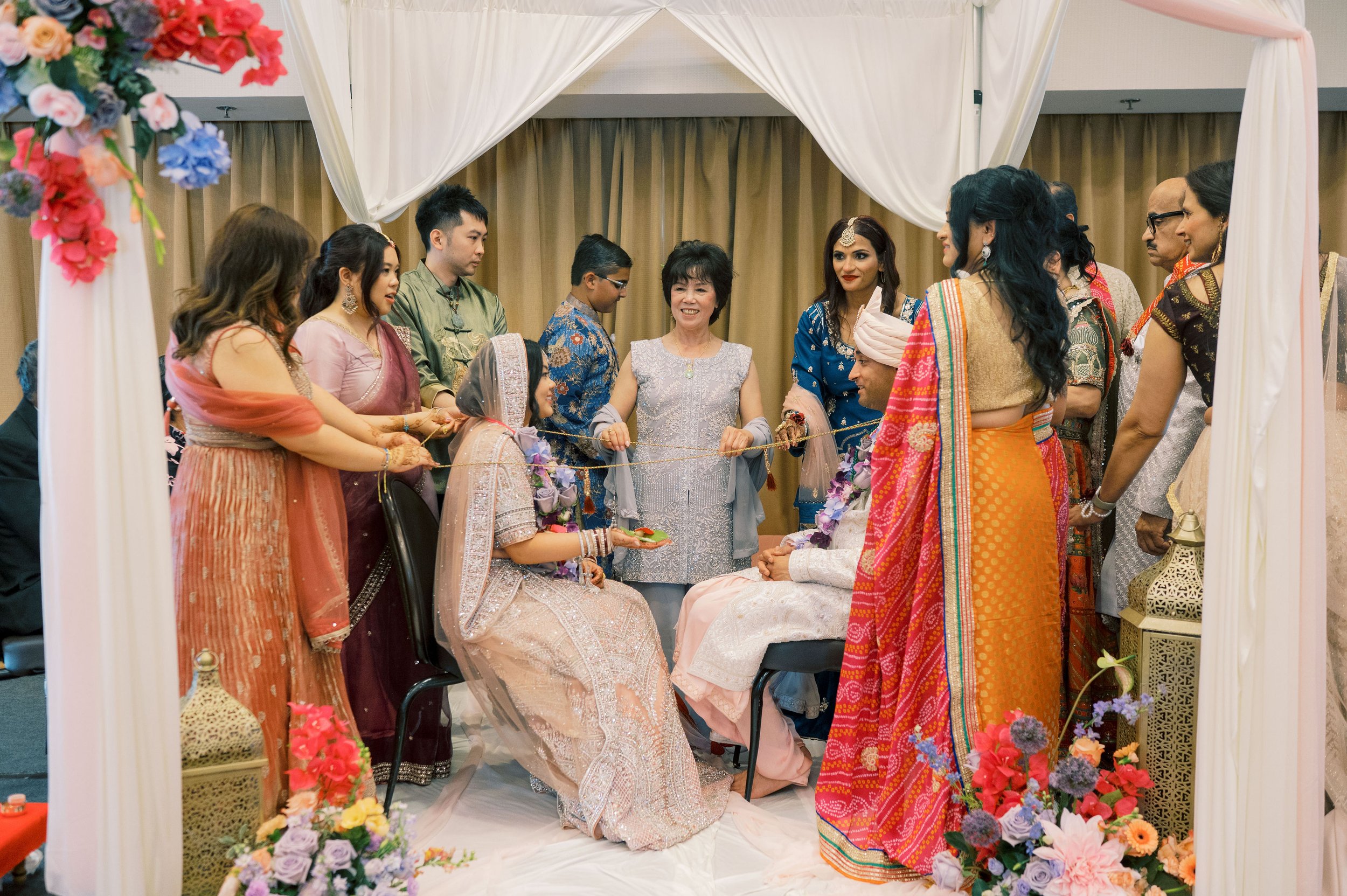 A multicultural group of people dressed in colorful traditional clothing gathered inside a decorated wedding arch with flowers. The scene appears to be a wedding or engagement ceremony.