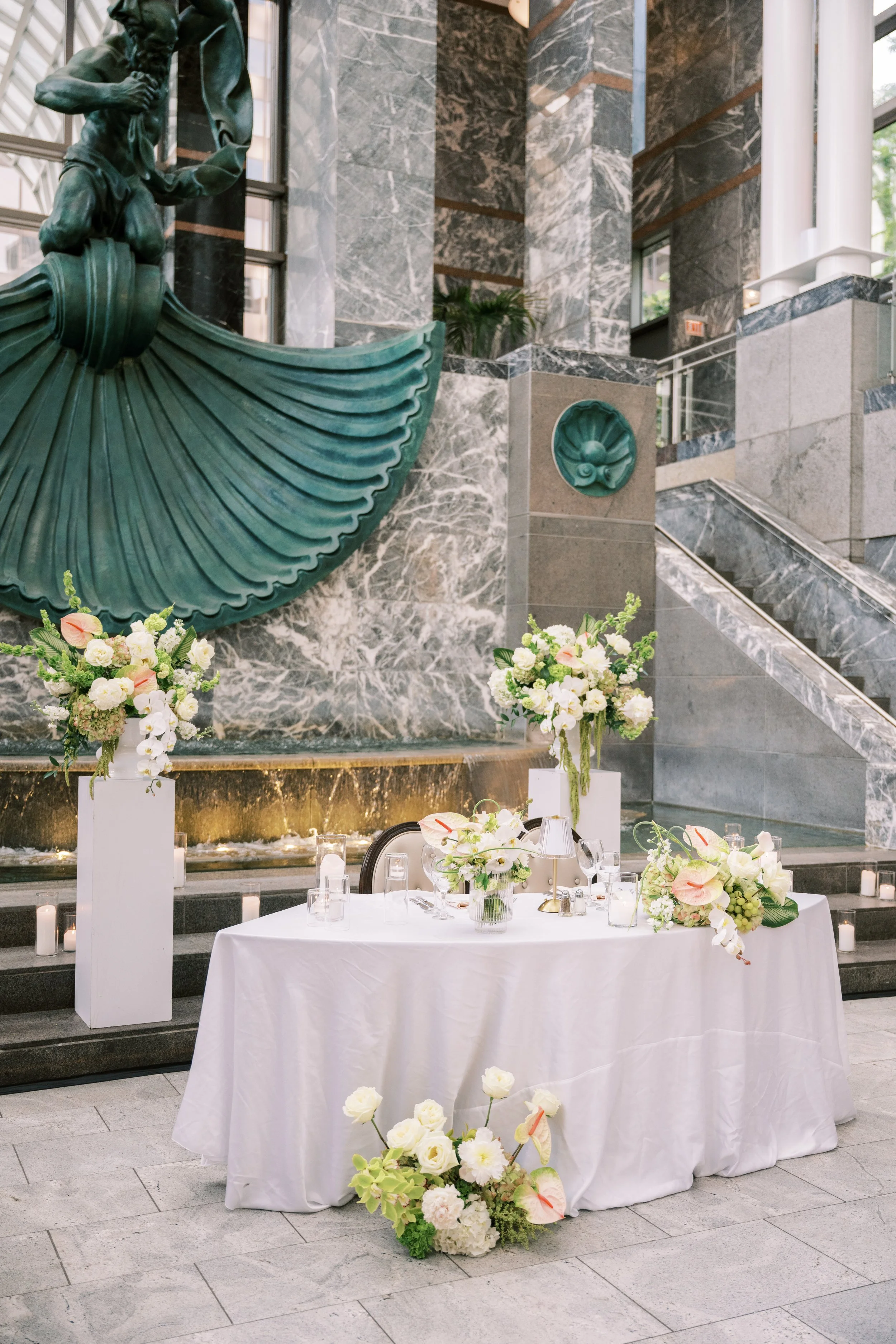 Elegant event setup with a table decorated with white tablecloth, floral centerpieces, and lit candles in a modern building with marble walls and large windows.