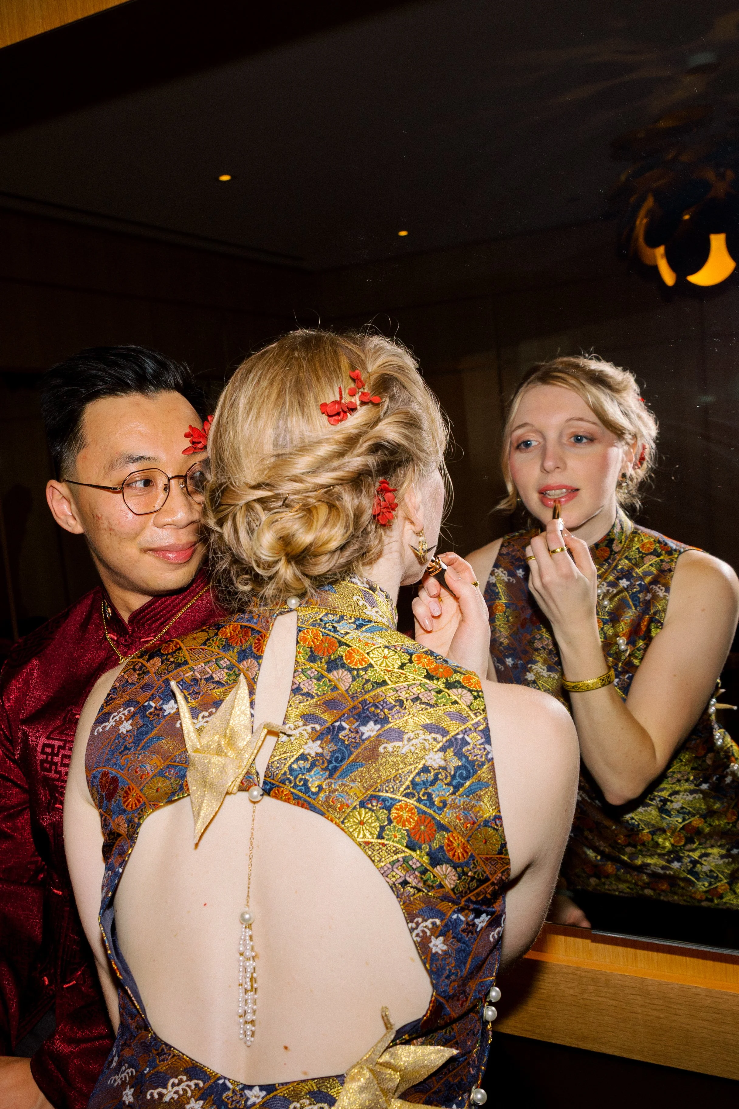 Young woman with blonde hair in an ornate, traditional Asian dress with floral and gold patterns, and decorative ornaments in her hair, applying makeup in front of a mirror. Two other people are partially visible nearby.