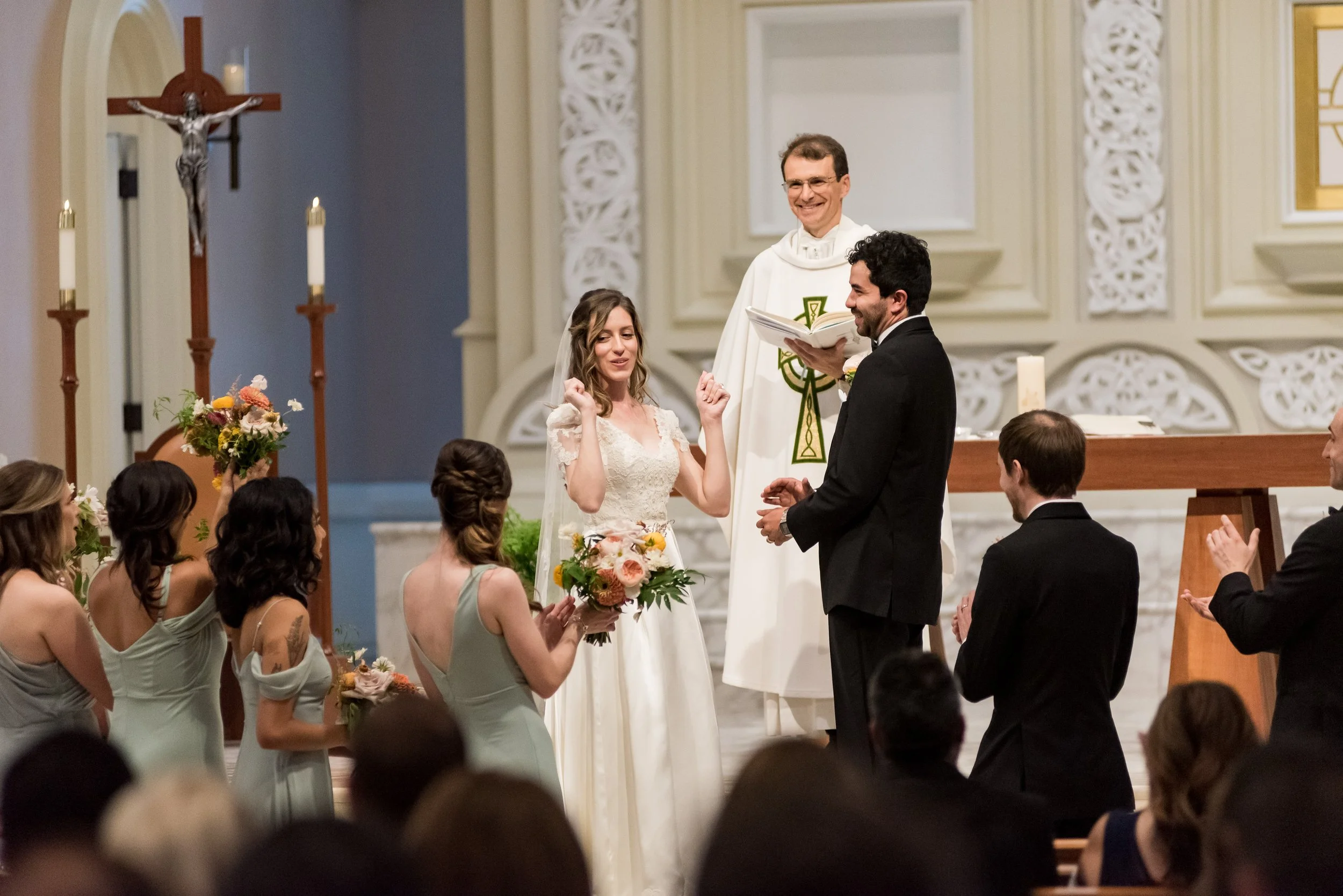 A wedding ceremony taking place in a church with a priest, the bride and groom standing at the altar surrounded by bridesmaids and groomsmen, flowers, candles, and a crucifix in the background.