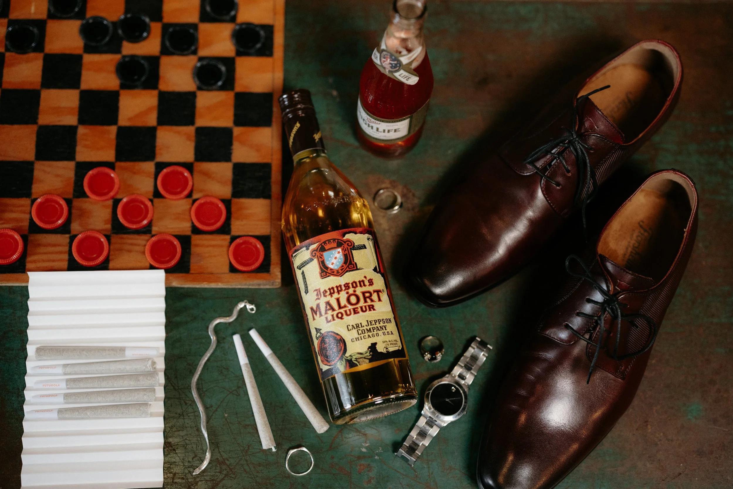 A collection of items including used checker pieces, two bottles of alcohol, a pair of brown leather shoes, a wristwatch, paper strips, and a white plastic object, arranged on a green surface.