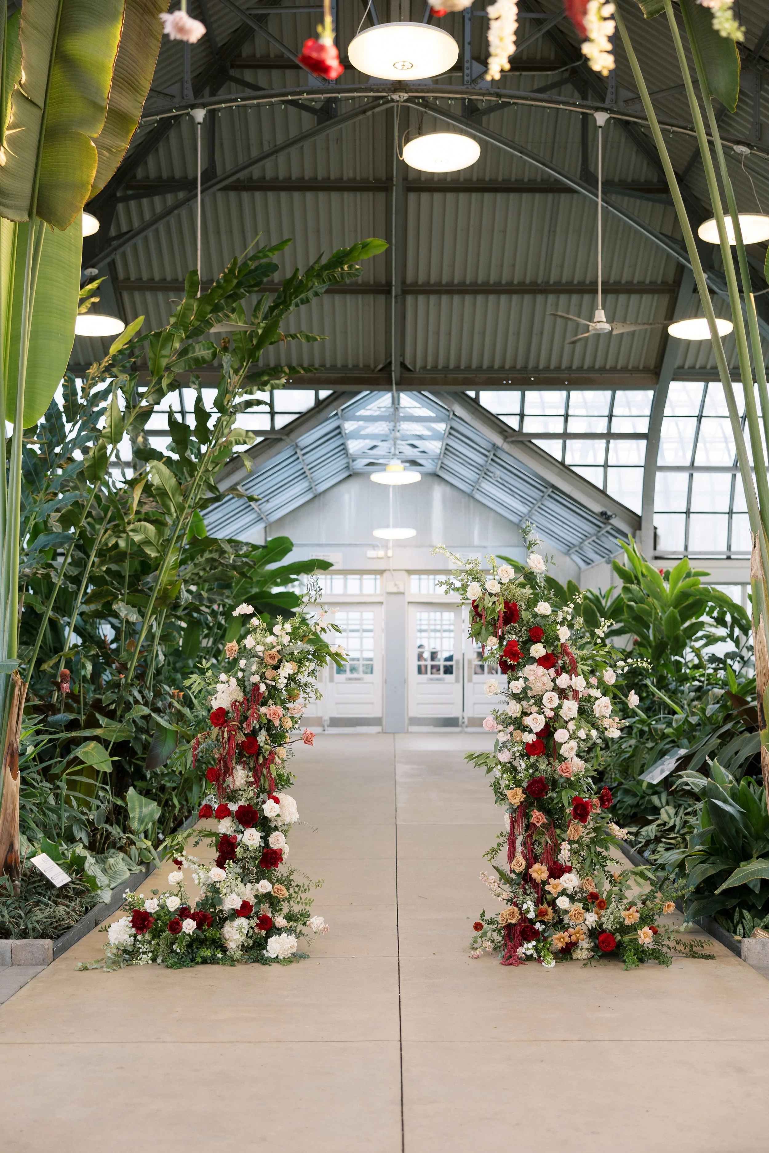 Indoor greenhouse or conservatory with large floral displays on either side of a wide walkway, plants on both sides, and a glass roof with metal framework, ceiling fans, and lights.