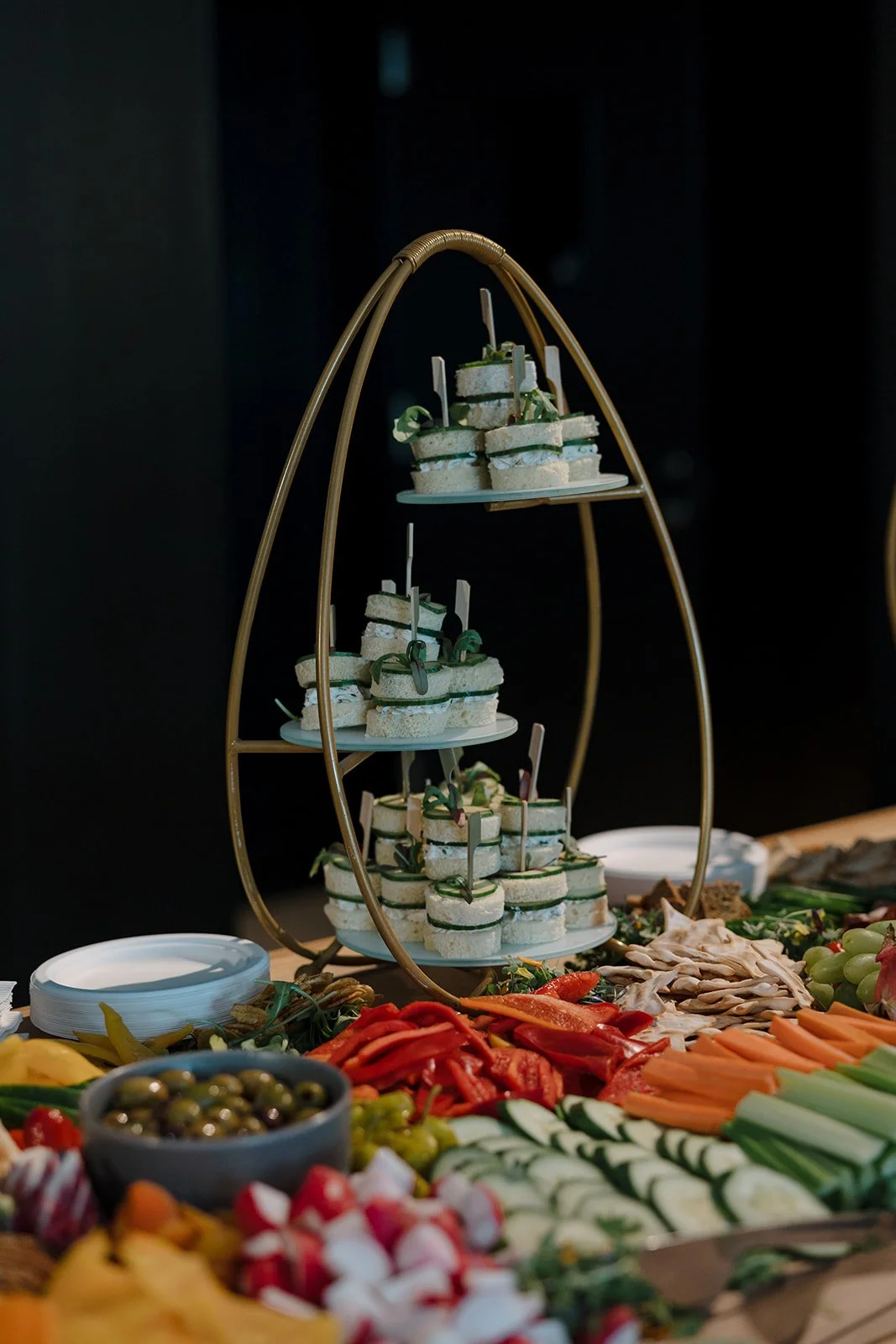 Tiered tray holding small sandwiches with green garnishes, surrounded by a variety of colorful vegetables, fruits, and sliced meats on a buffet table.
