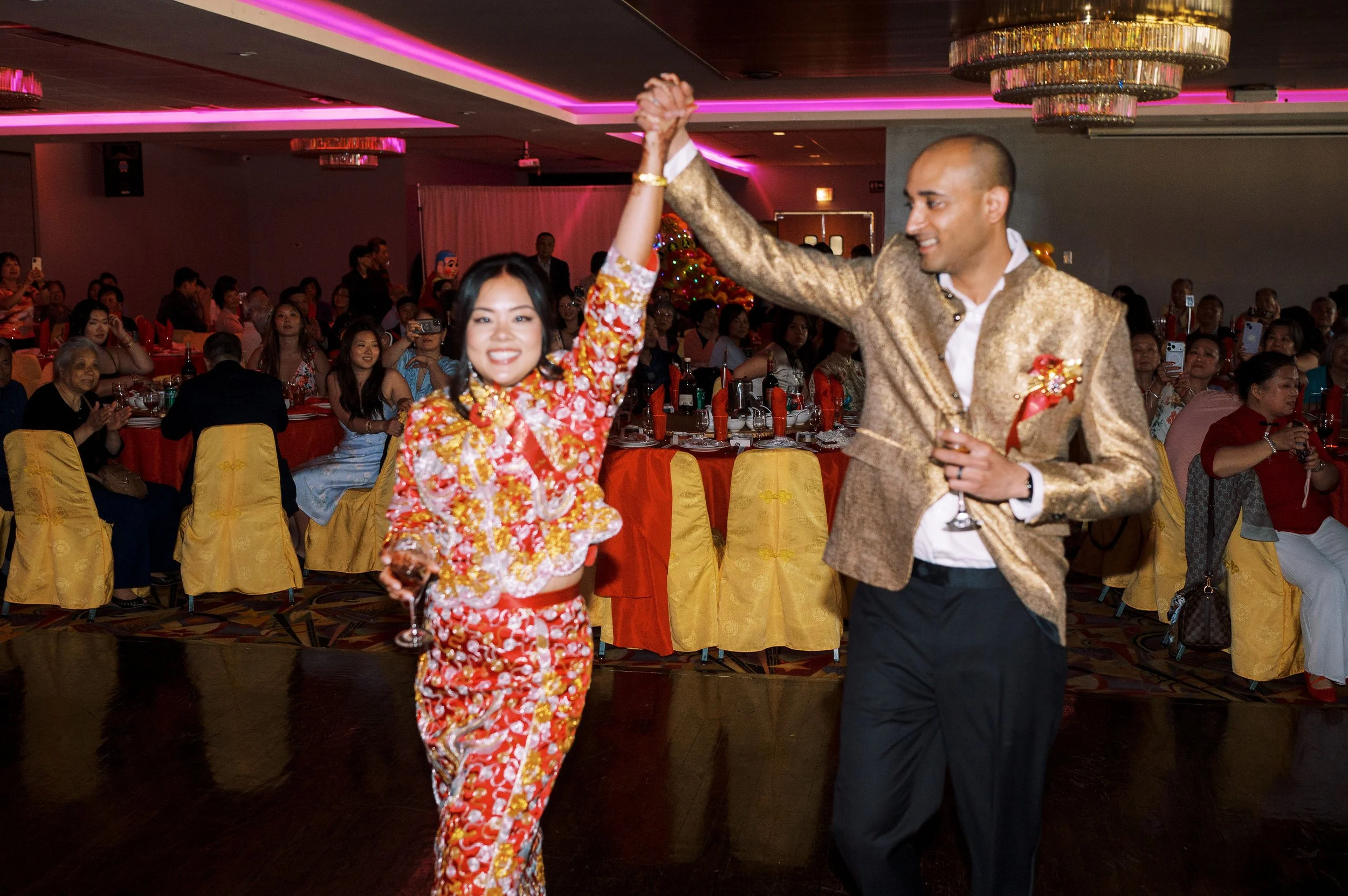 A man and woman dancing together at a festive celebration, with the woman wearing a colorful traditional dress and the man in a gold blazer, surrounded by seated guests.