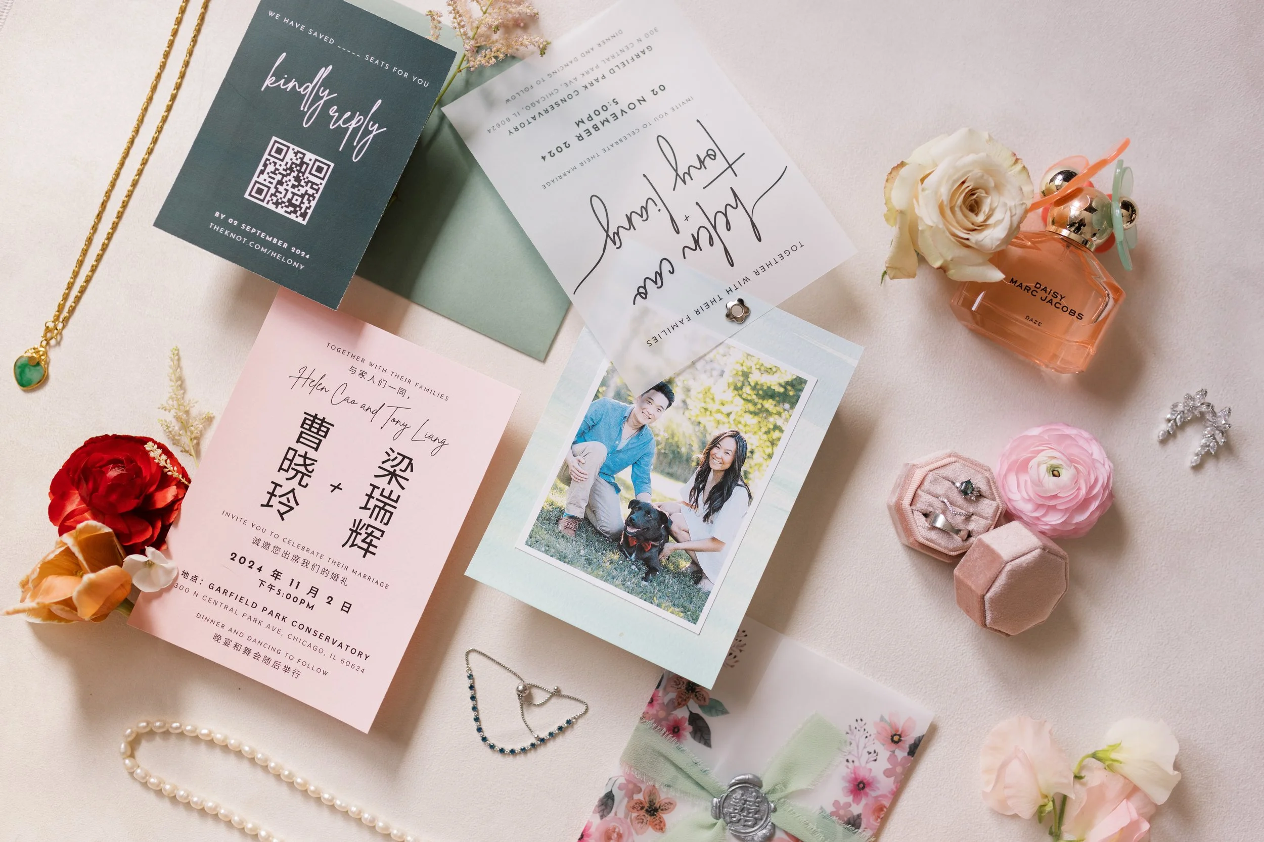 Wedding invitation cards, jewelry including a pearl necklace and a silver bracelet, a fragrance bottle labeled 'Daisy Marc Jacobs,' flowers, and decorative items are arranged on a white surface for a wedding celebration.