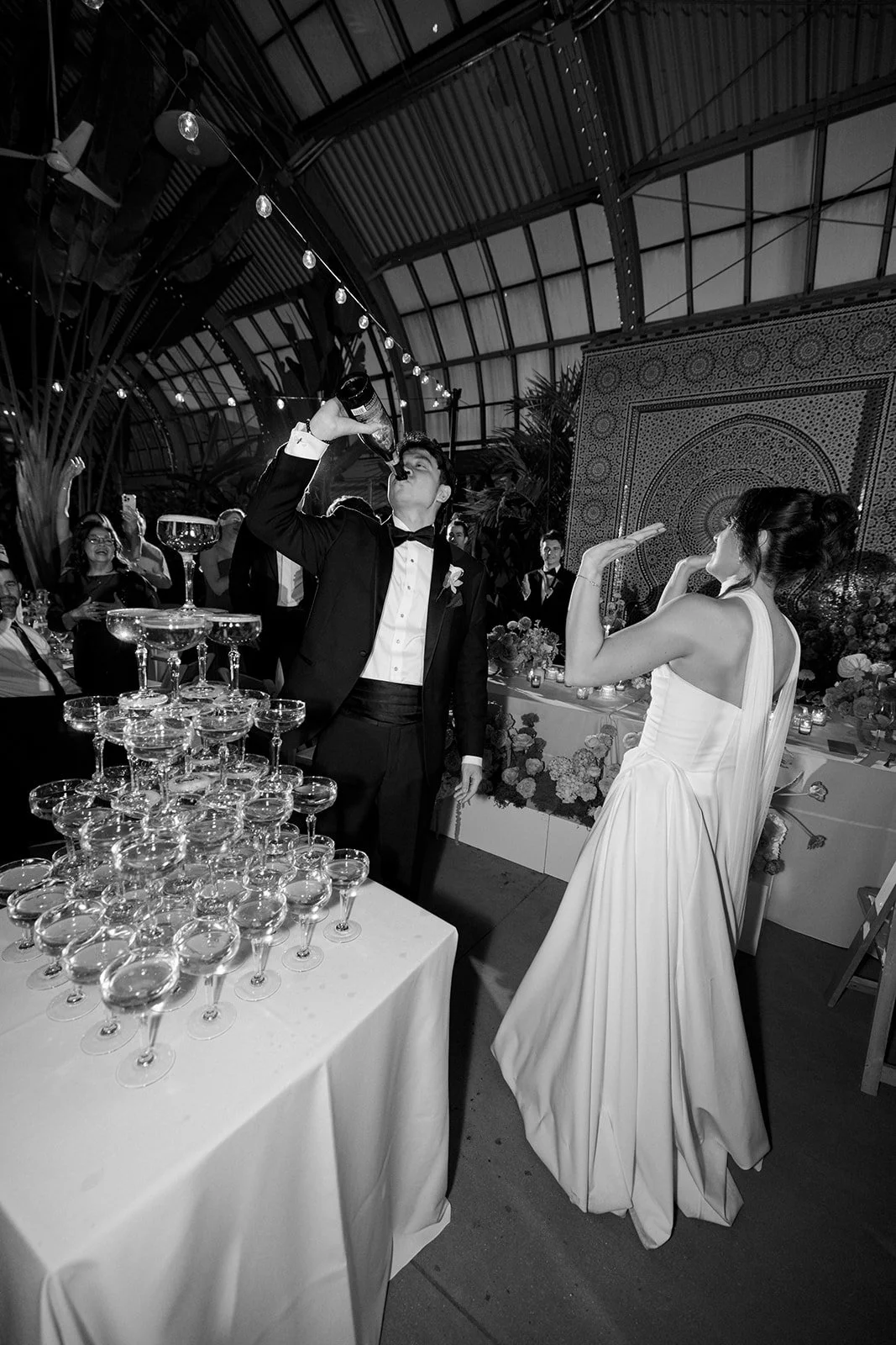 A black and white wedding reception scene with a groom in a tuxedo drinking from a bottle, and a bride in a wedding dress gesturing with her hand. Guests are seated at tables decorated with flowers and candles, with a pyramid of champagne glasses in 