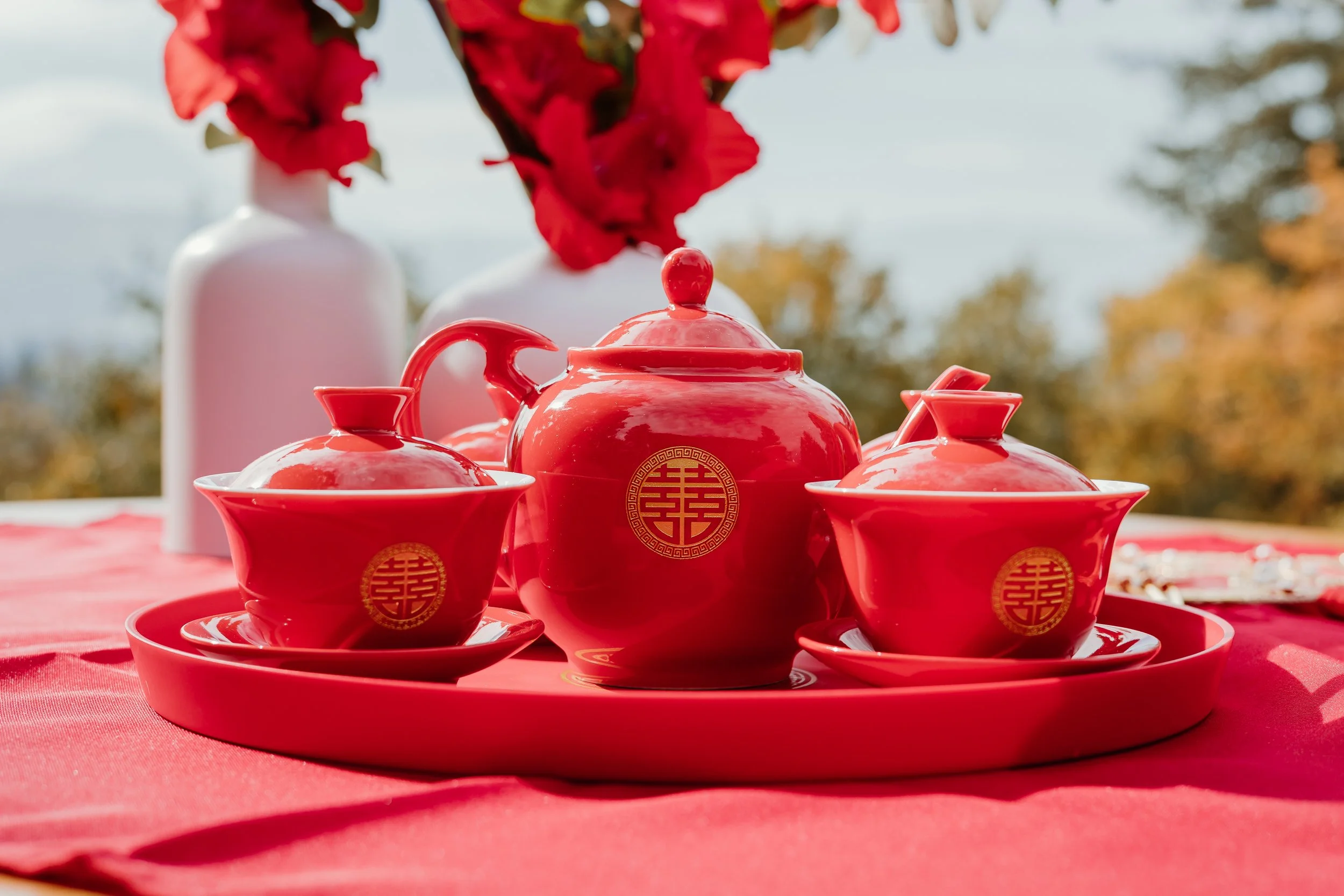 Red tea set with cups, teapot, and tray, decorated with gold symbols, placed on a red tablecloth outdoors.