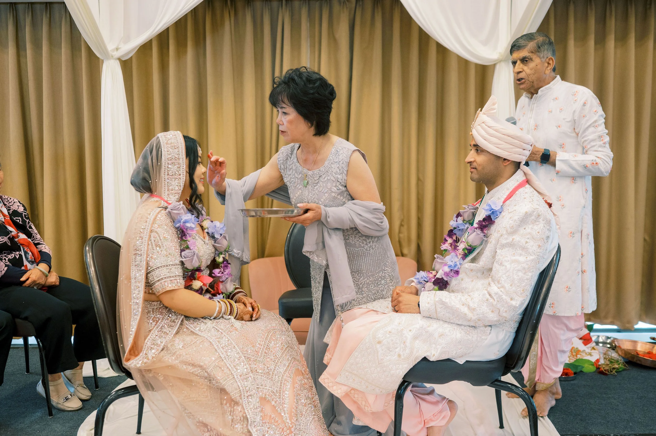 A traditional wedding ceremony with the bride and groom dressed in ornate attire seated, with family members around them. An elderly woman is applying sindoor or similar to the bride's forehead, while an elderly man holds a microphone nearby. The bac