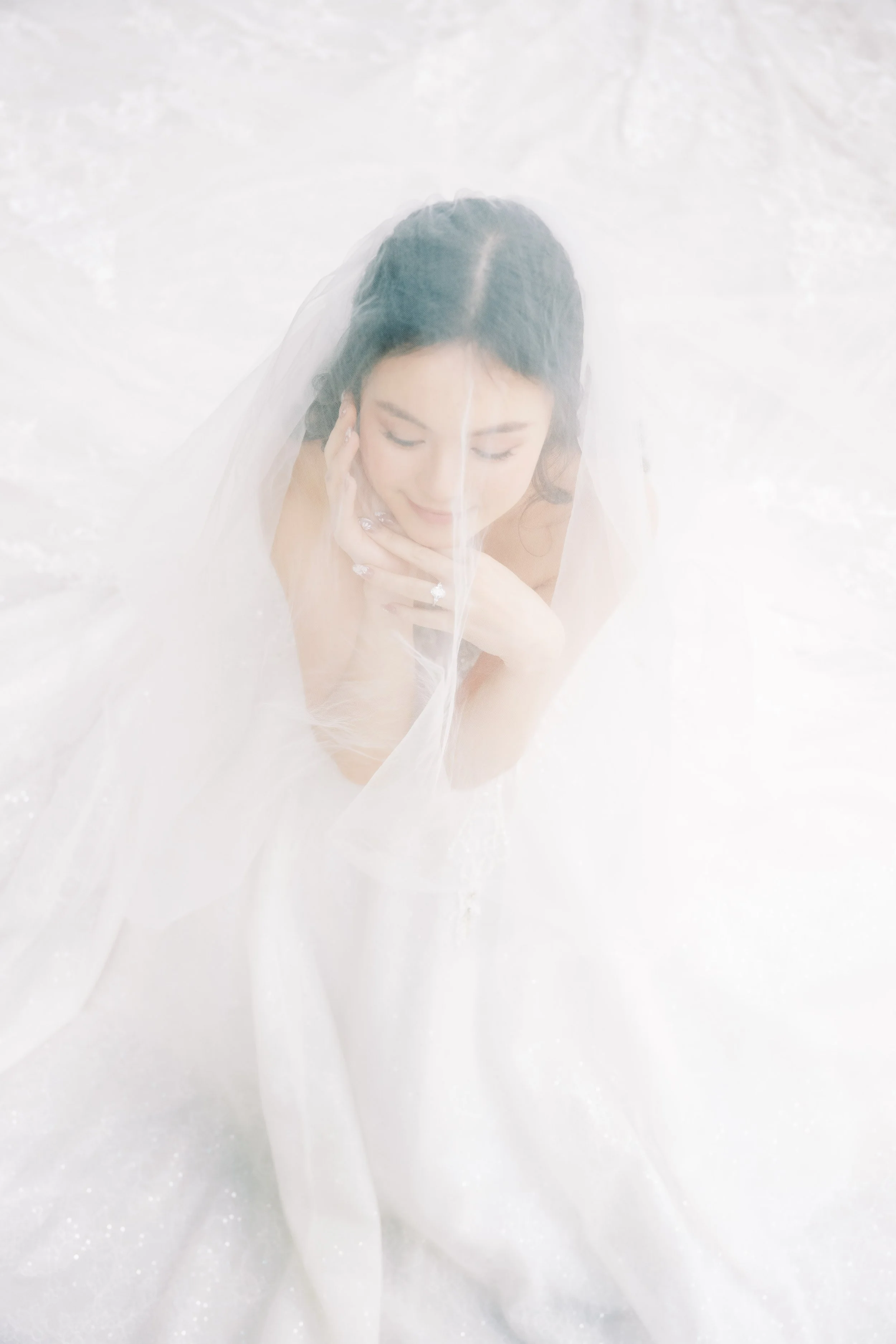 A woman with dark hair and closed eyes wearing a white wedding dress and veil, smiling with her hands touching her face, standing in a snowy or frosty environment.