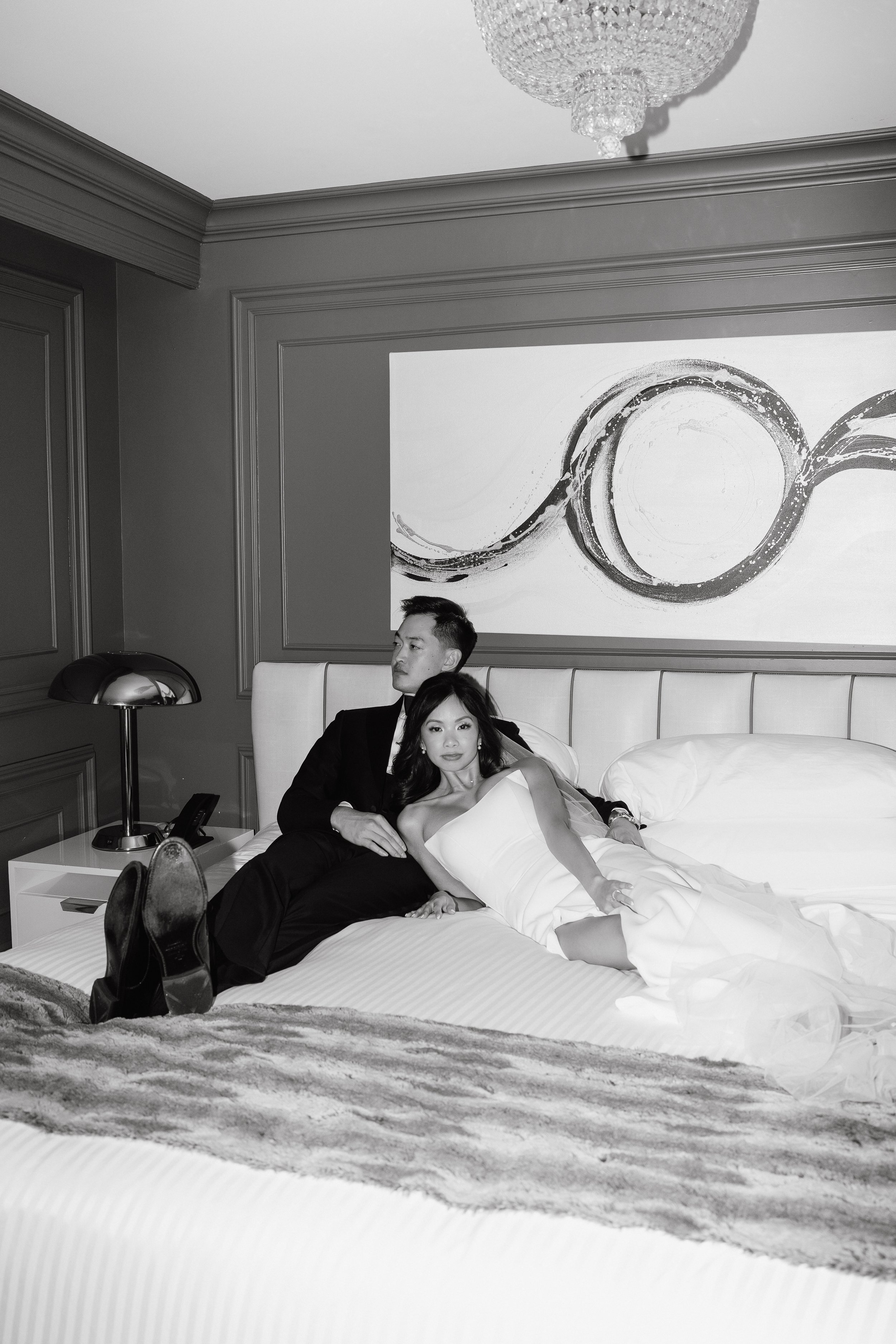 A black-and-white photo of a man and woman in formal attire sitting on a bed in a bedroom. The woman is lying back on the bed while the man is sitting, leaning against the headboard. There is artwork on the wall behind them and a lamp on the bedside 