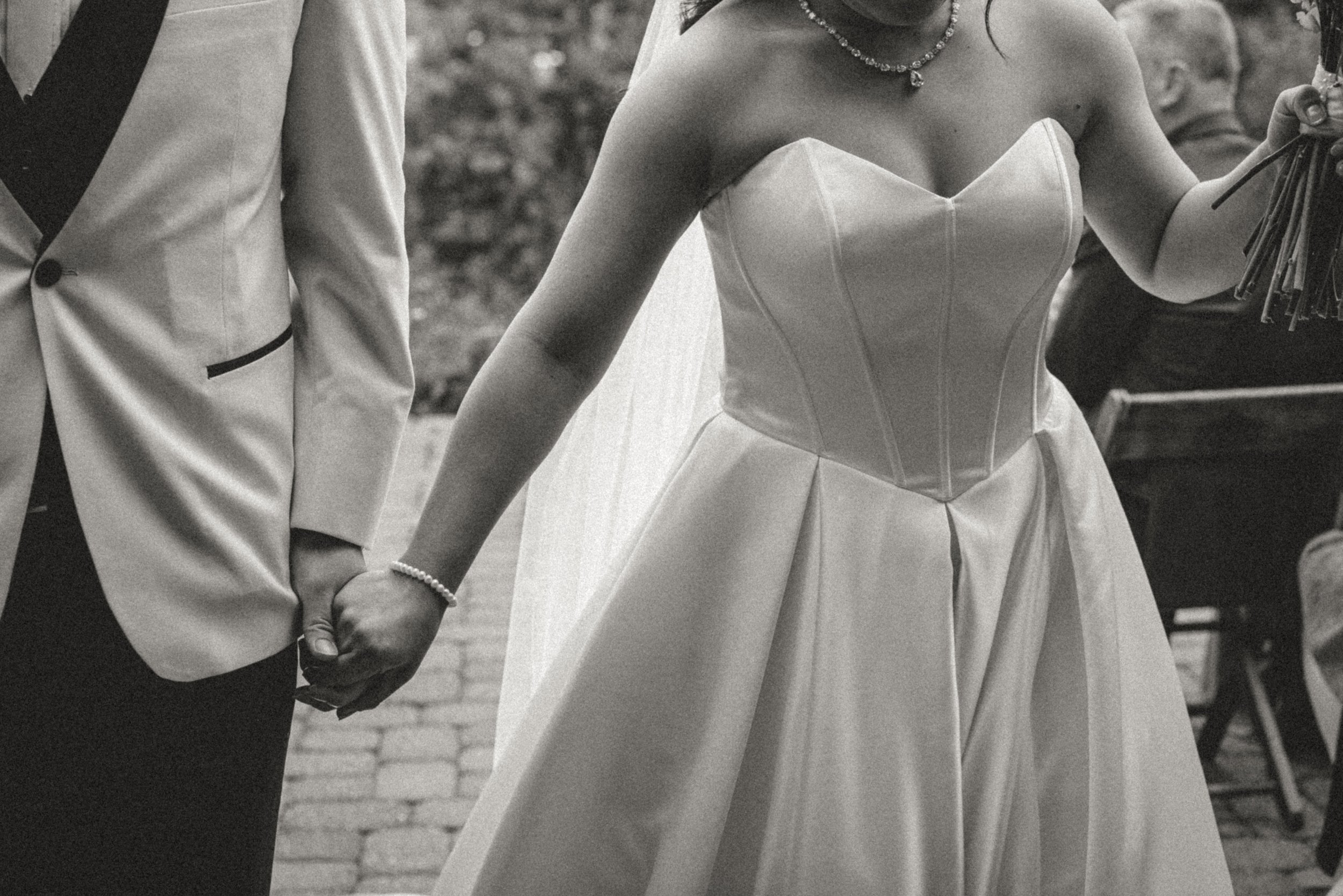 A black and white photo of a couple holding hands during a wedding ceremony. The bride wears a strapless gown and a necklace, and is holding a bouquet. The groom wears a light-colored suit with dark trim and a boutonniere. They are standing outdoors 
