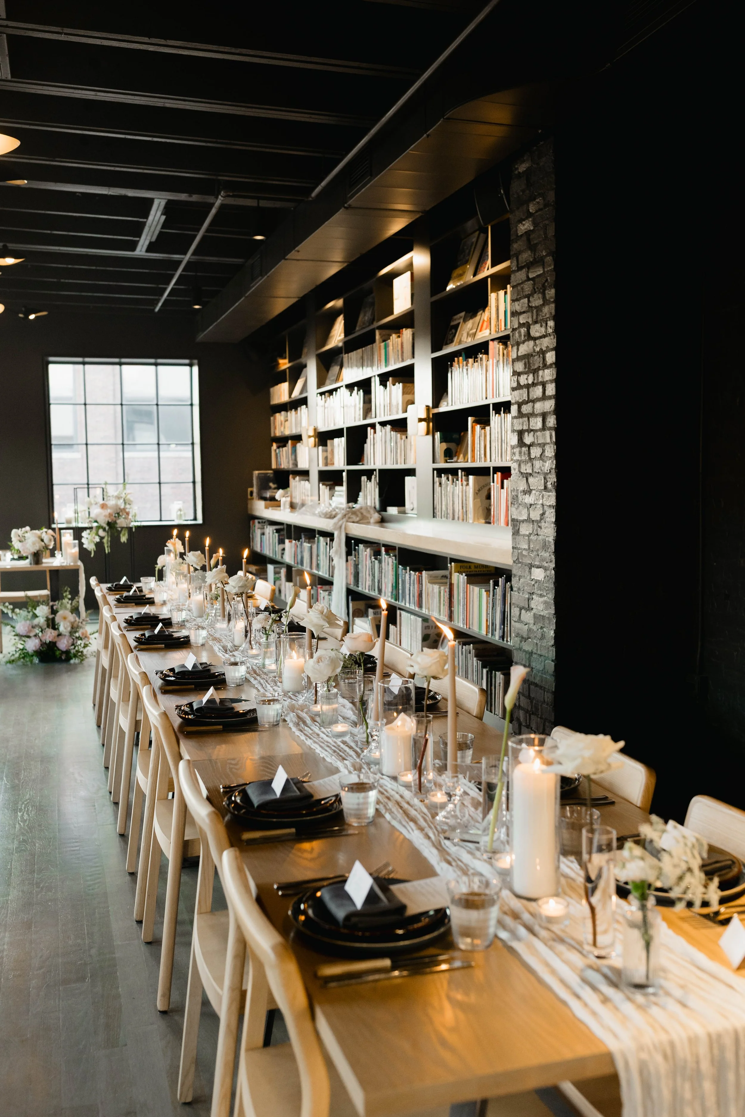 Long wooden dining table set with black dinnerware, white napkins, and lit candles for a formal event in a modern, industrial-style room with exposed brick wall and bookshelves.