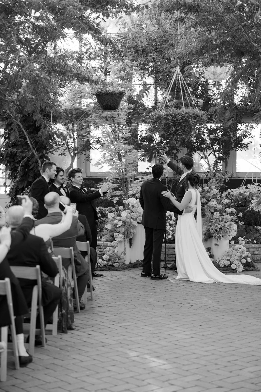 A black and white photo of a wedding ceremony with a bride and groom standing together. The couple is exchanging vows or rings, surrounded by bridesmaids and groomsmen. Guests are seated, some taking photos. The ceremony is held outdoors, decorated w
