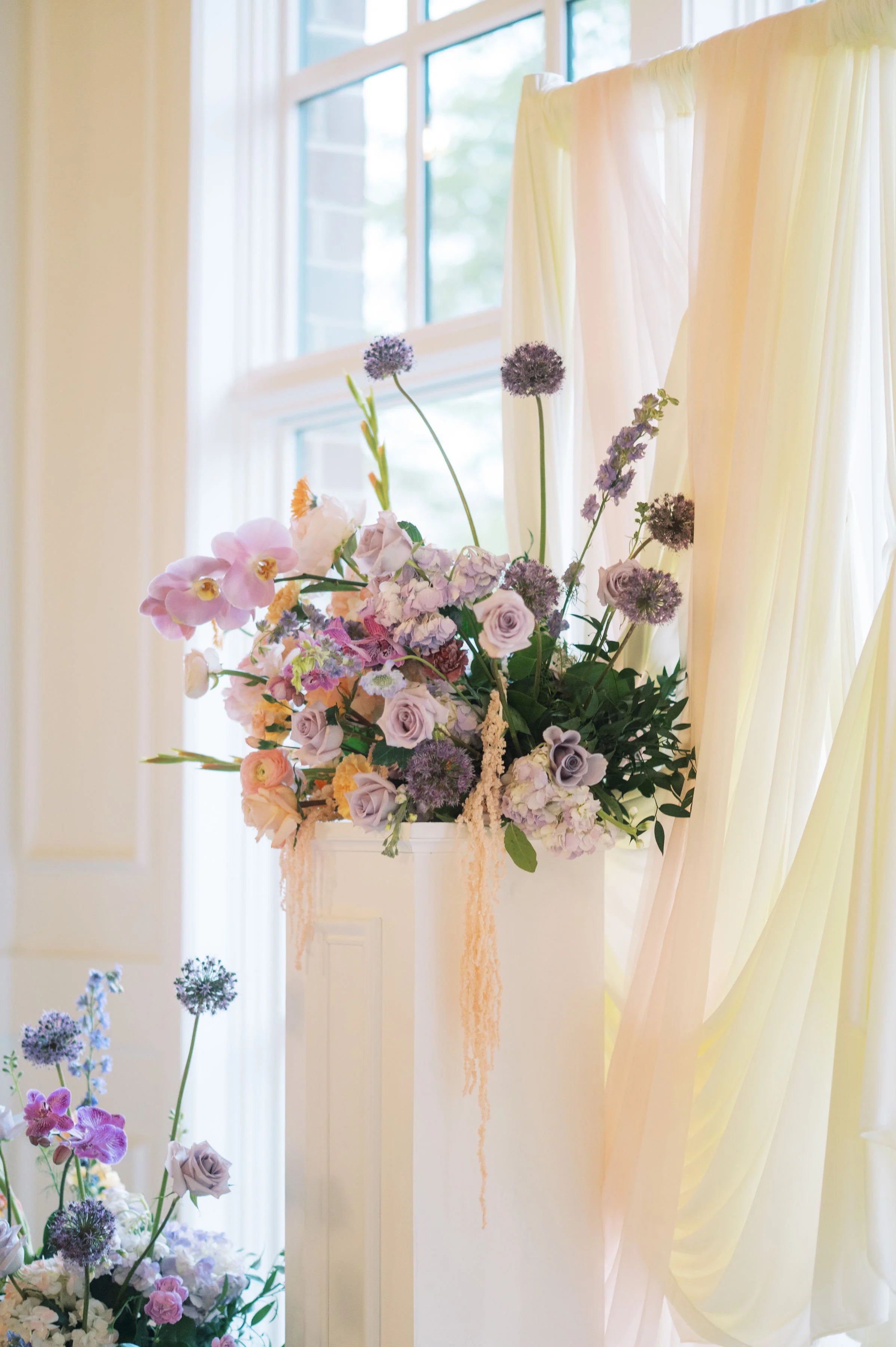 A floral arrangement with purple, pink, and white flowers, hanging on a white wall next to a window with cream-colored curtains, in a bright room.