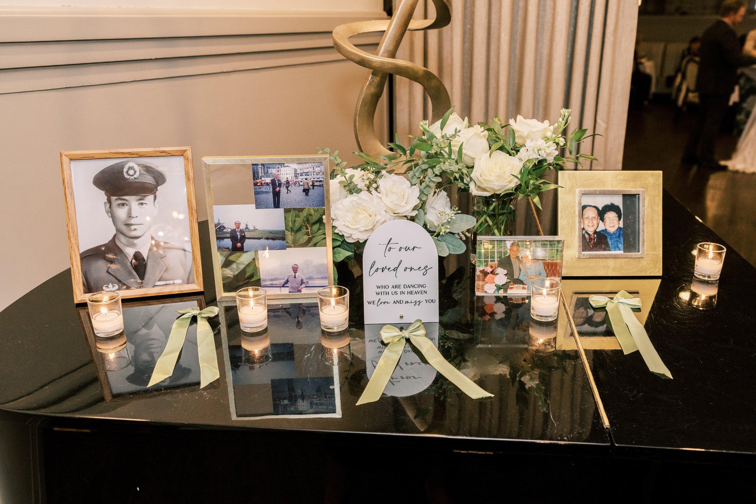 Memorial table with black and white portrait of a young man in military uniform, surrounded by framed photographs of various people, white flowers, lit candles, and a note that reads 'to our loved ones who are dancing with us in heaven, we love and m
