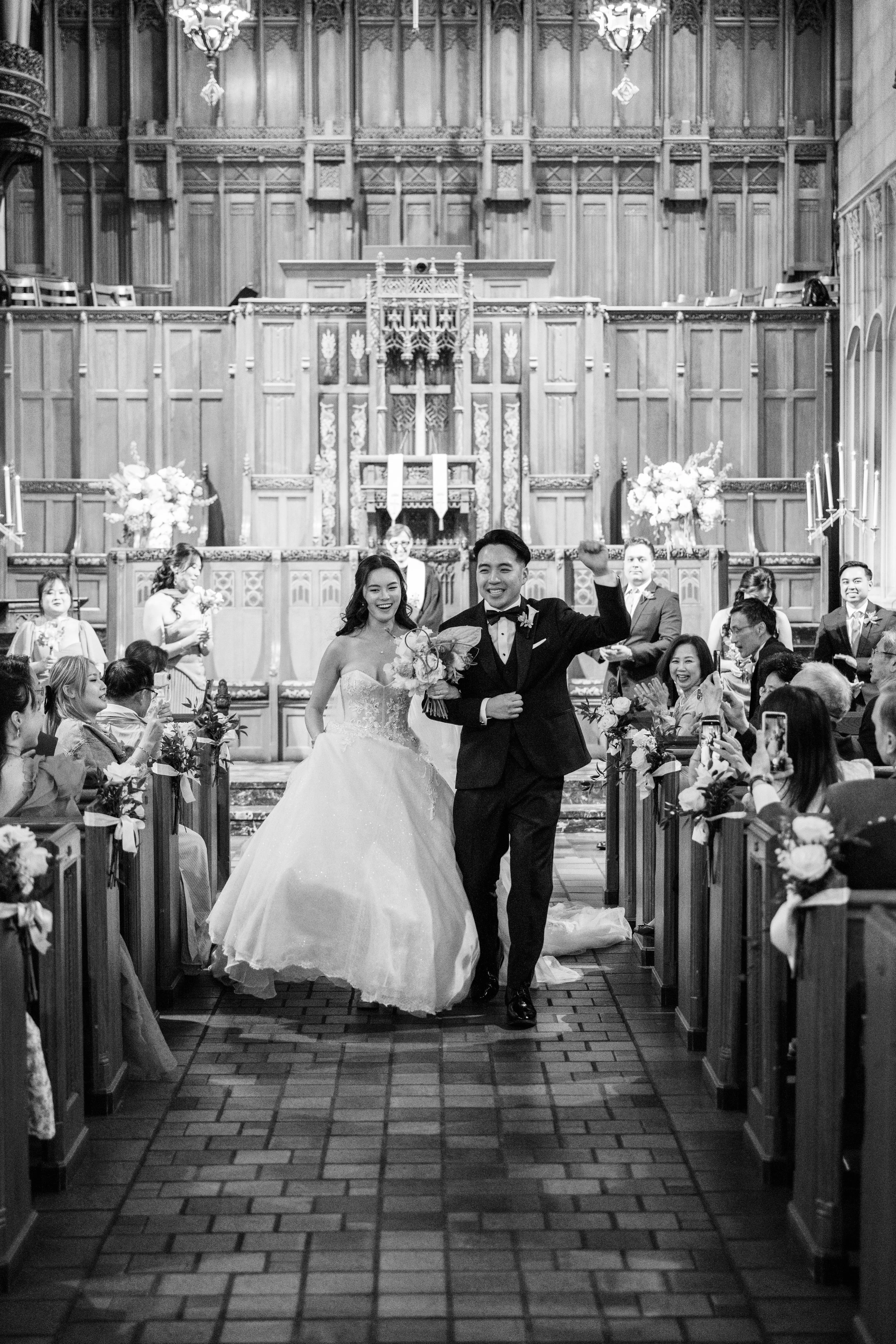 A bride and groom walking down the aisle in a church, surrounded by seated guests, with flowers and candles along the sides.