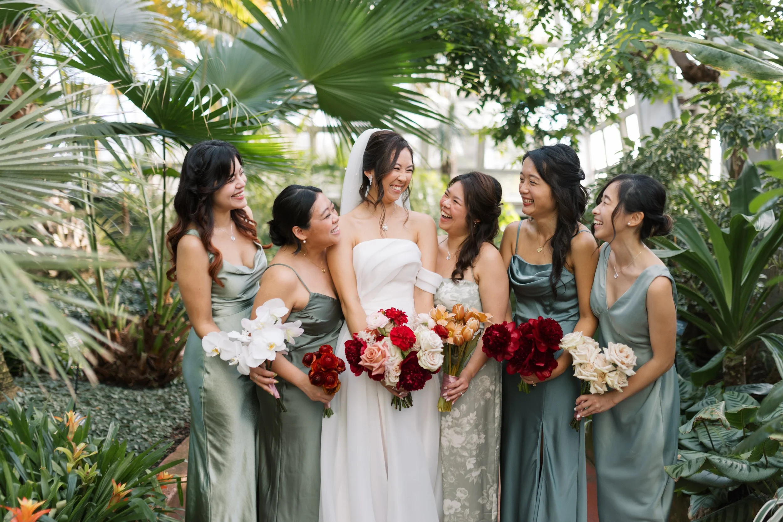 A bride in a white wedding gown surrounded by five women in matching satin dresses holding bouquets of red, pink, white, and orange flowers, all smiling and enjoying a moment together in a lush, green conservatory.