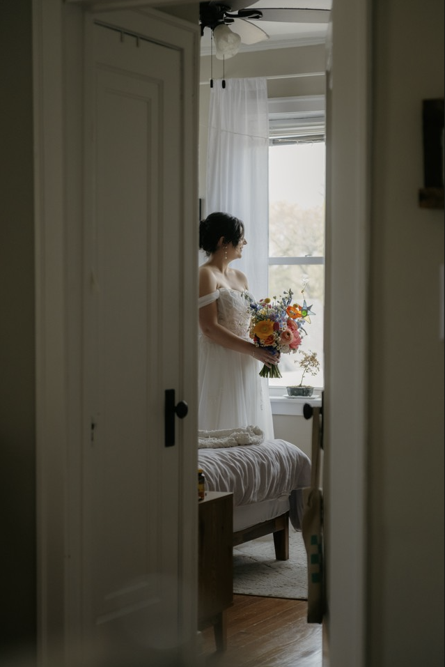 A bride in a white dress holding a colorful bouquet of flowers, standing near a window in a softly lit room, seen through a doorway.