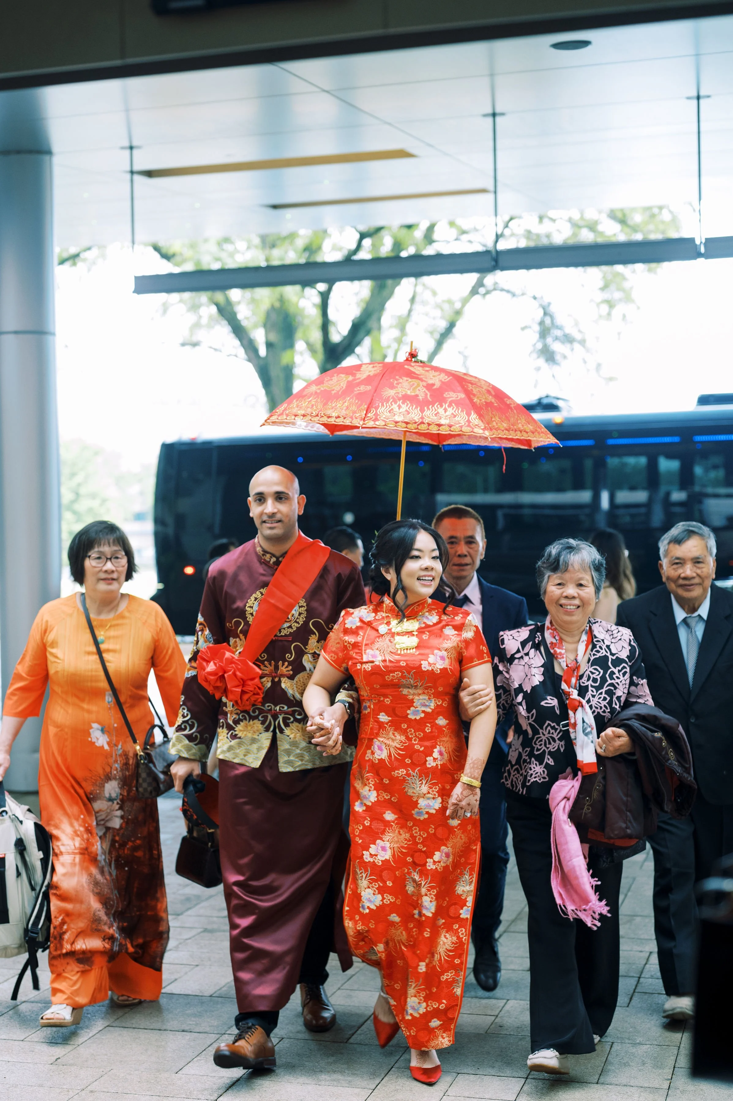 People dressed in traditional Asian attire walking with a person holding a red umbrella, under a modern building entrance.
