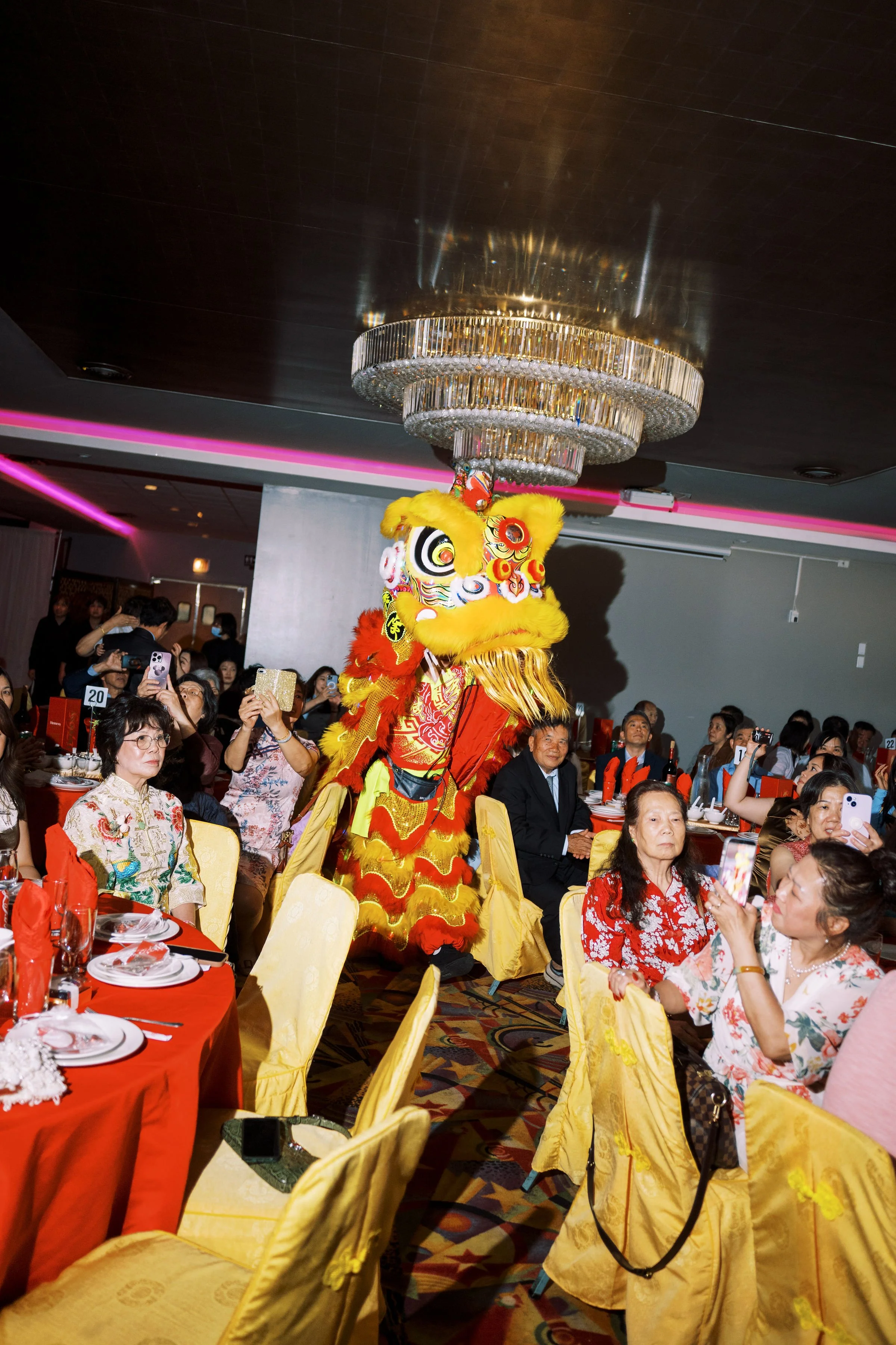 A Chinese lion dance performer in a yellow and red lion costume performing in front of an audience in a decorated banquet hall, with a large crystal chandelier hanging from a dark ceiling.