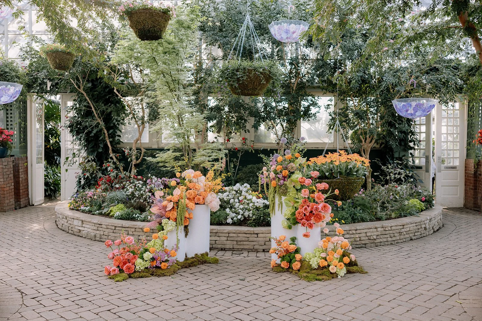 Inside a greenhouse filled with hanging flower baskets, potted plants, and colorful blooming flowers arranged in white vases and garden beds.