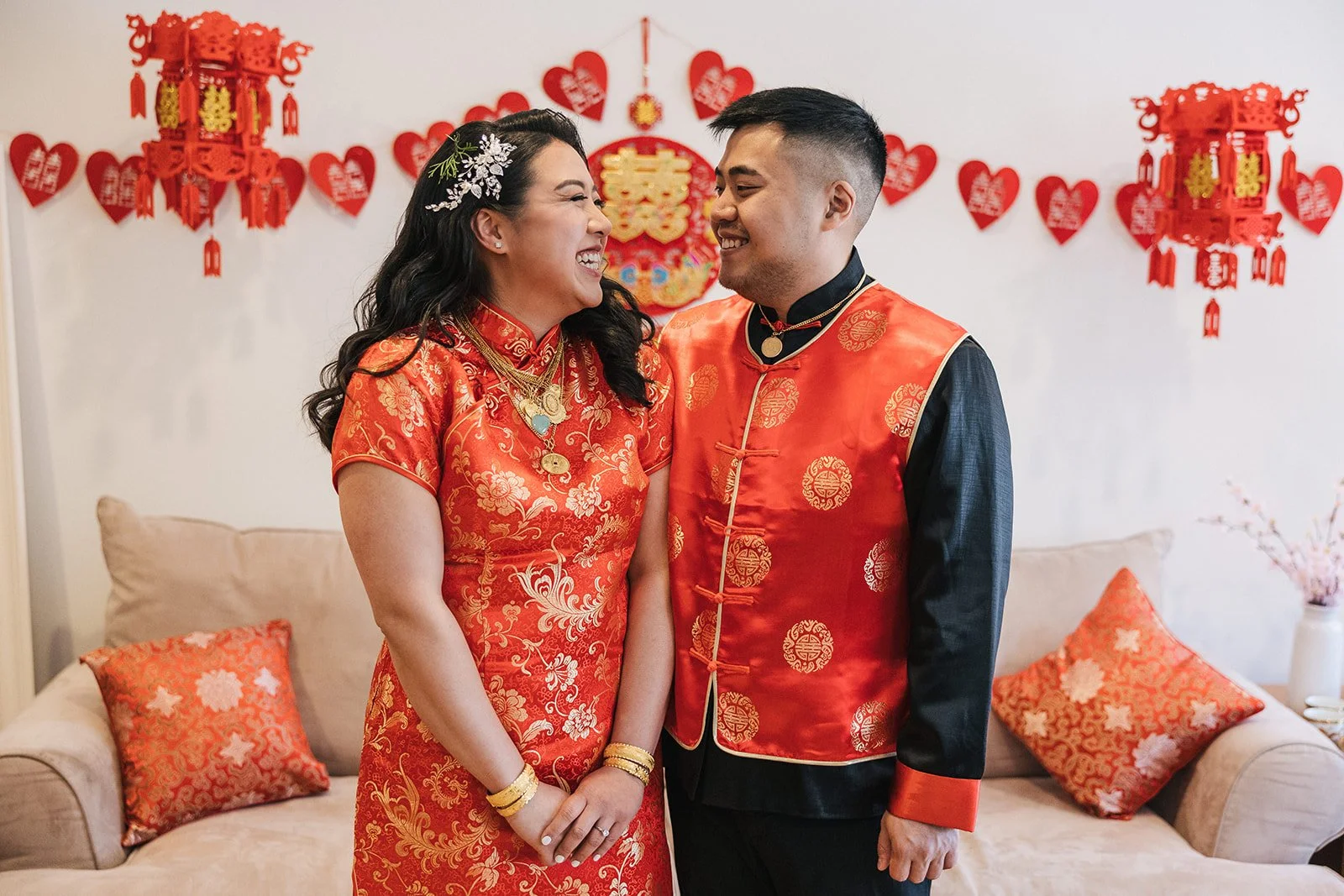 A smiling couple dressed in traditional Chinese wedding attire, standing close together in a decorated room with red and gold decorations, including hanging lanterns and heart-shaped ornaments on the wall.