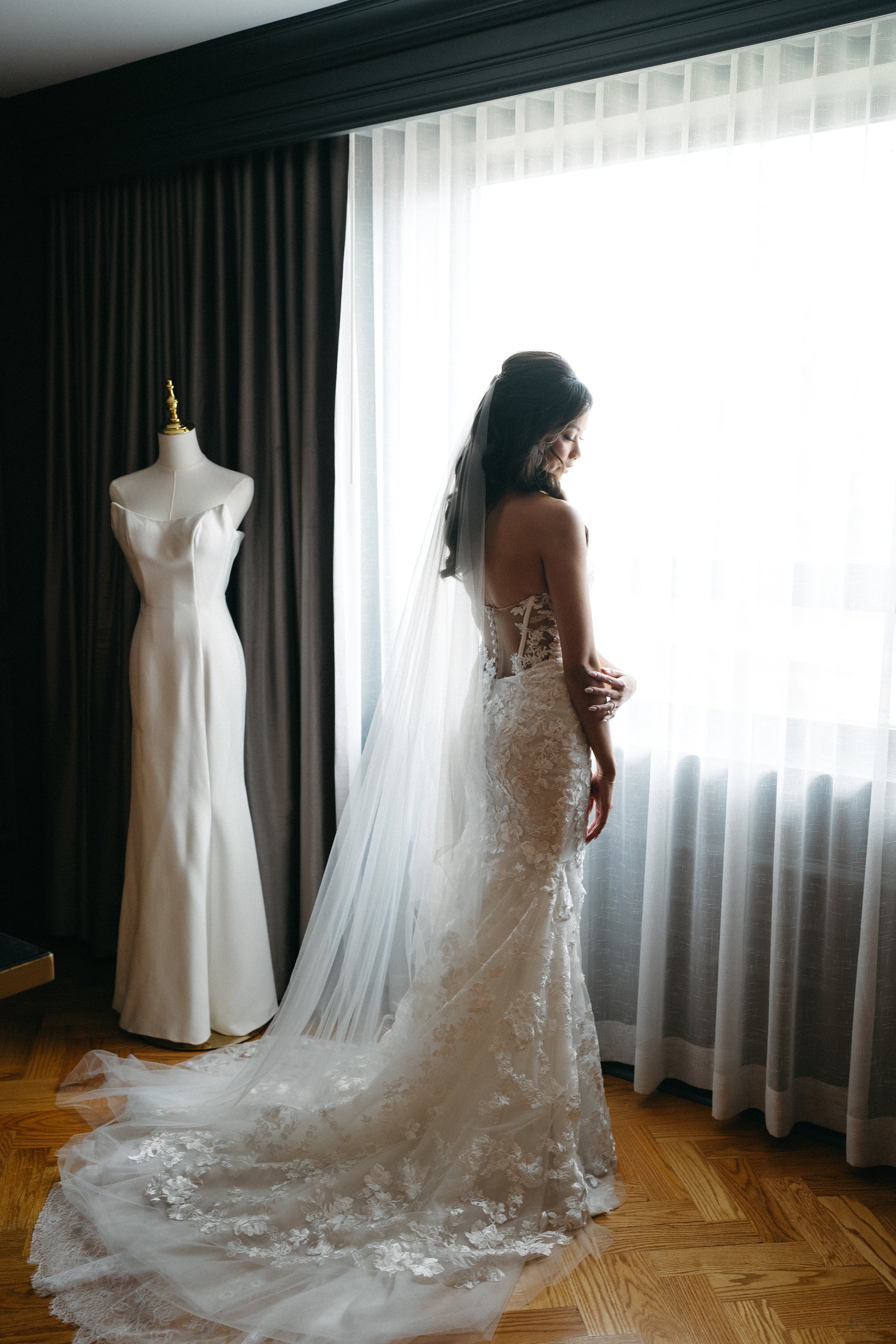 A bride in a lace wedding gown with a long train and veil stands near large window with sheer curtains, next to an empty mannequin dressed in a white gown, in a dimly lit room.
