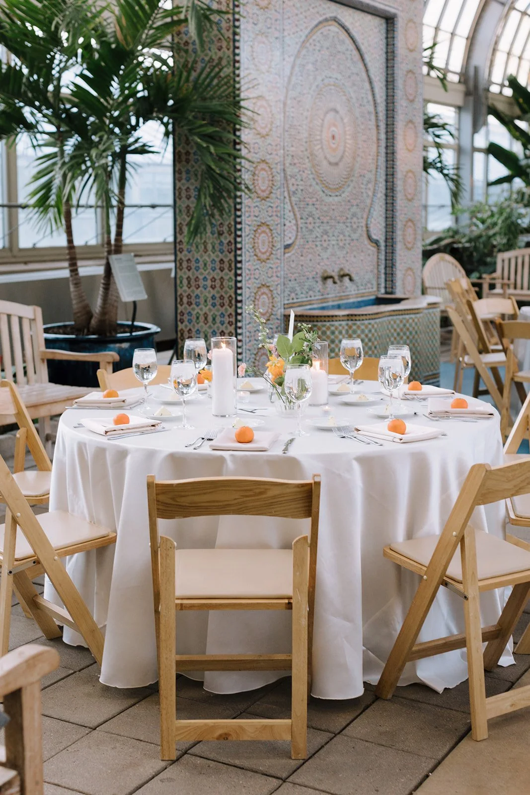A round banquet table with white tablecloth, set with water glasses, silverware, white napkins, and small peach-colored decorations. The centerpiece includes candles and fresh flowers. The background features green plants, a mosaic fountain, and seat