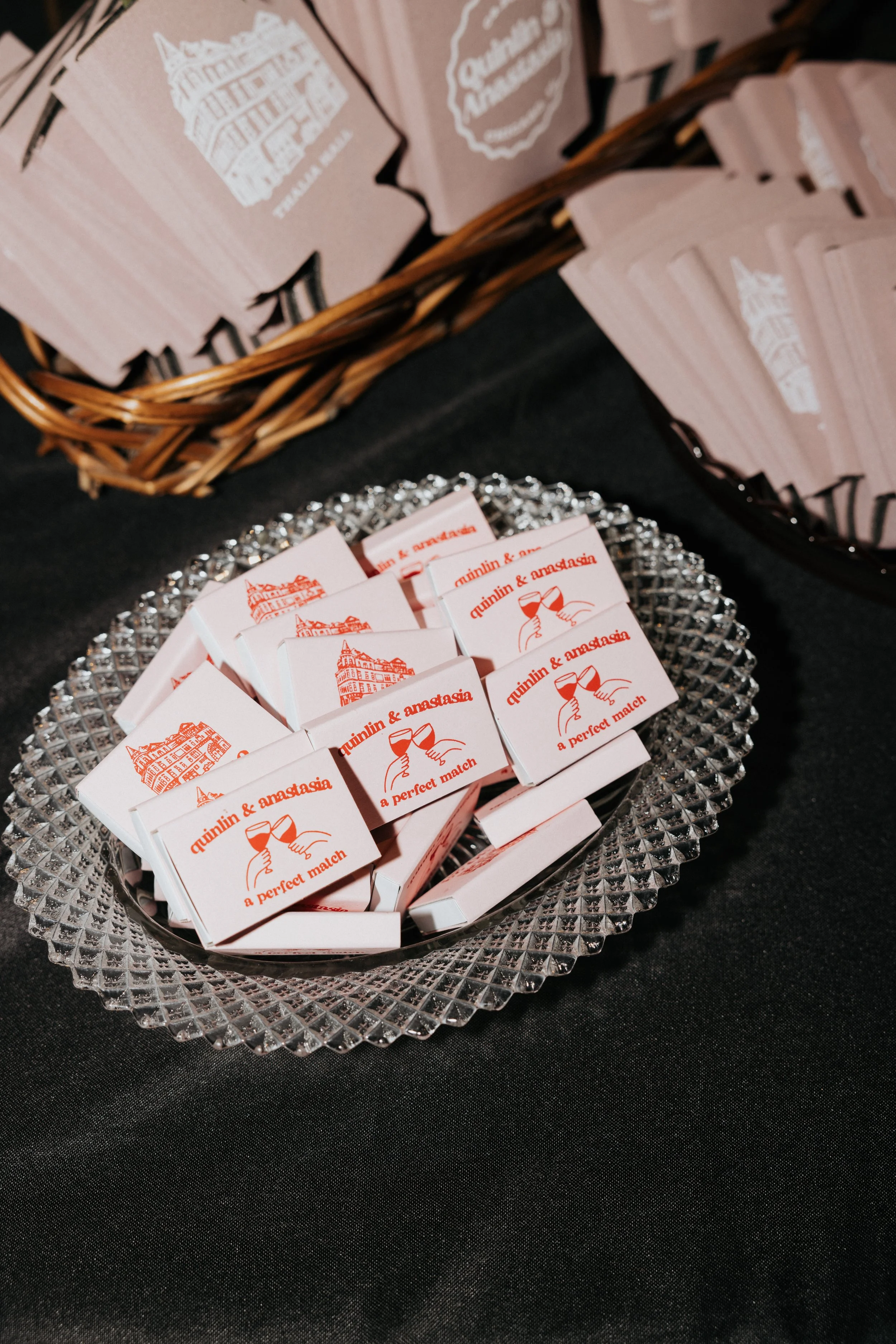 A glass dish filled with small paper matchboxes that have red and white designs, including an illustration of two hands holding wine glasses and the text "quilin & anastasia" and "a perfect match." In the background, there is a basket filled with pin