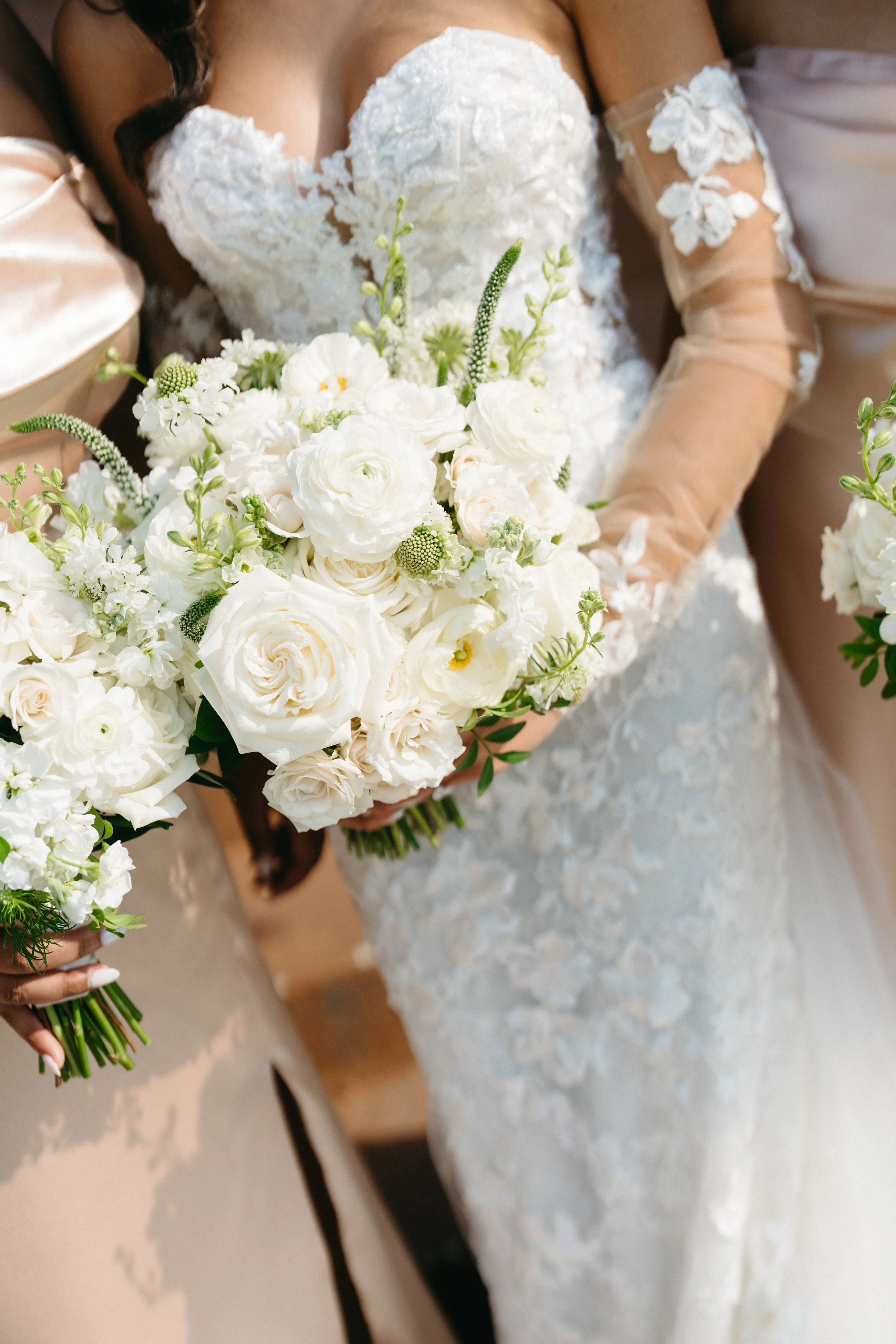 Bride in a white lace wedding dress holding a bouquet of white flowers, standing beside her partner in a beige suit.