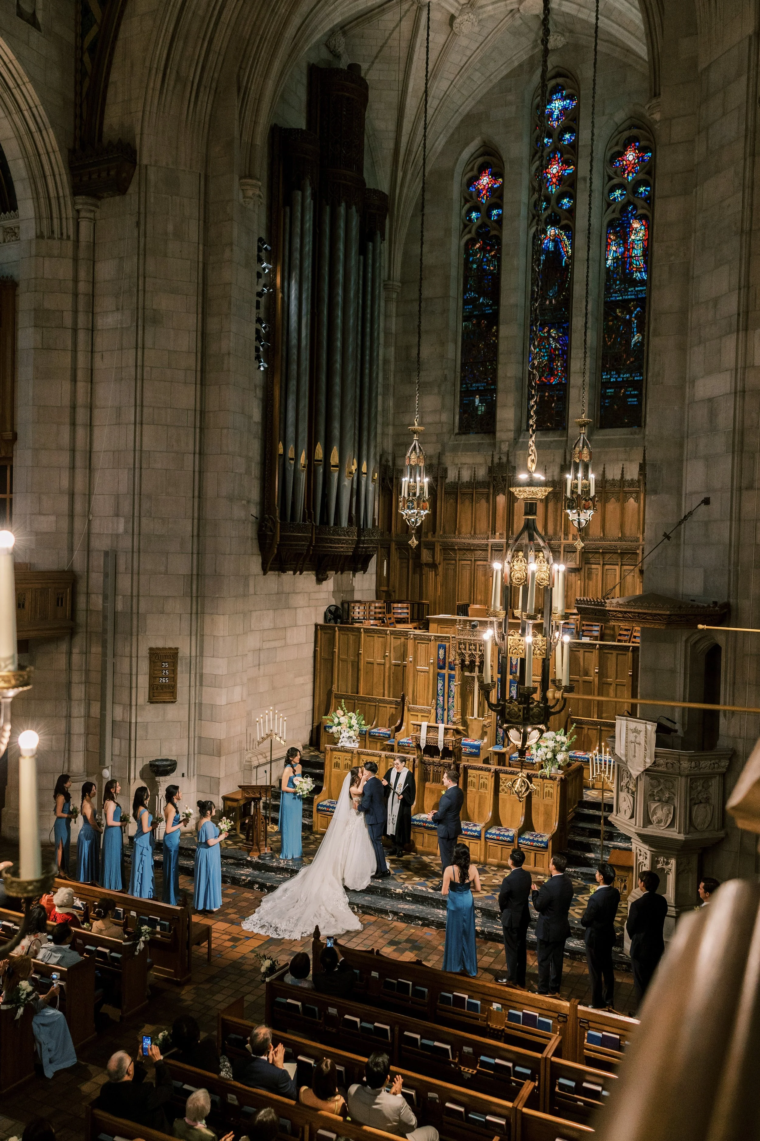 A wedding ceremony taking place inside a church with high vaulted ceilings, stained glass windows, and a large pipe organ. The bride and groom are standing at the altar with the officiant, surrounded by bridesmaids in blue dresses and groomsmen in da