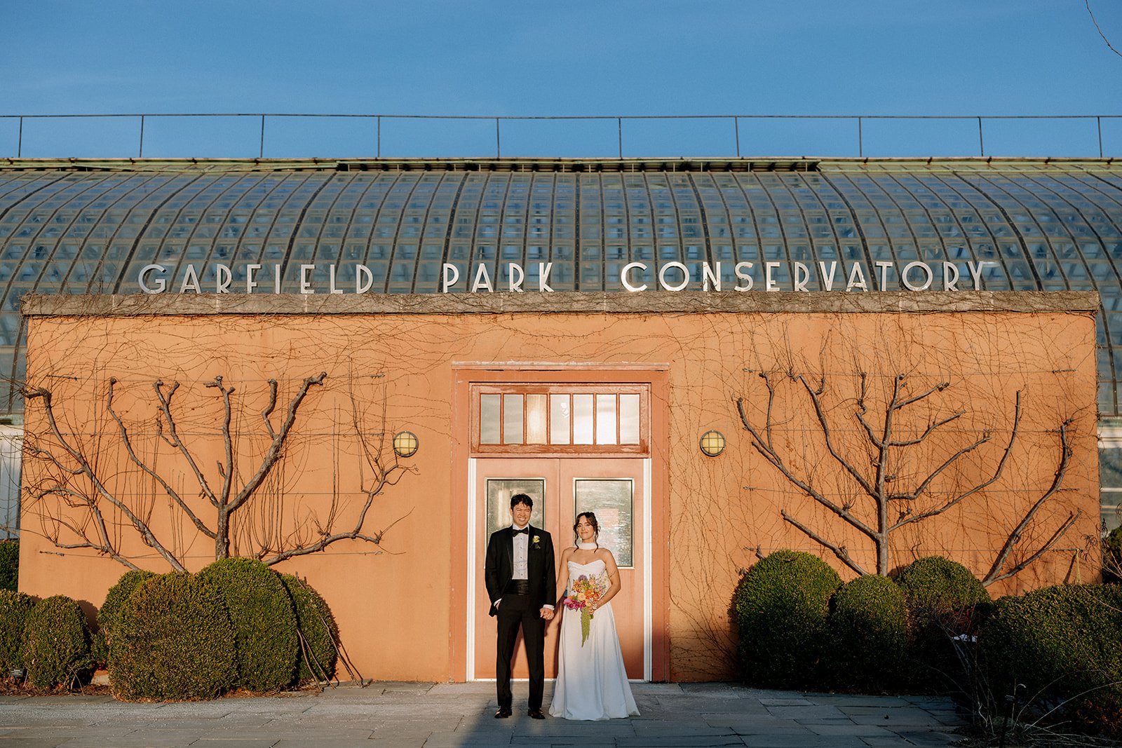 A bride and groom stand in front of a building labeled Garfield Park Conservatory, with leafless trees and bushes on either side.