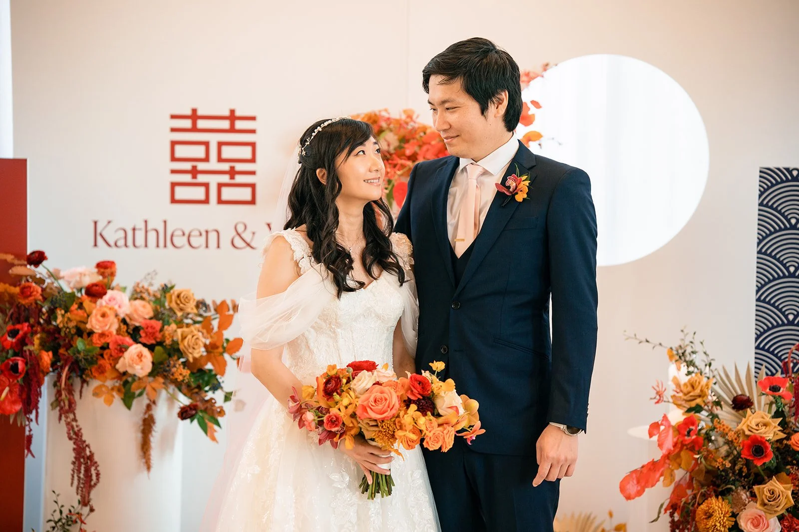 A bride and groom at their wedding ceremony, standing close and gazing at each other, with the bride holding a colorful bouquet in front of them. The background features floral decorations and a sign with Japanese characters and part of the names Kat
