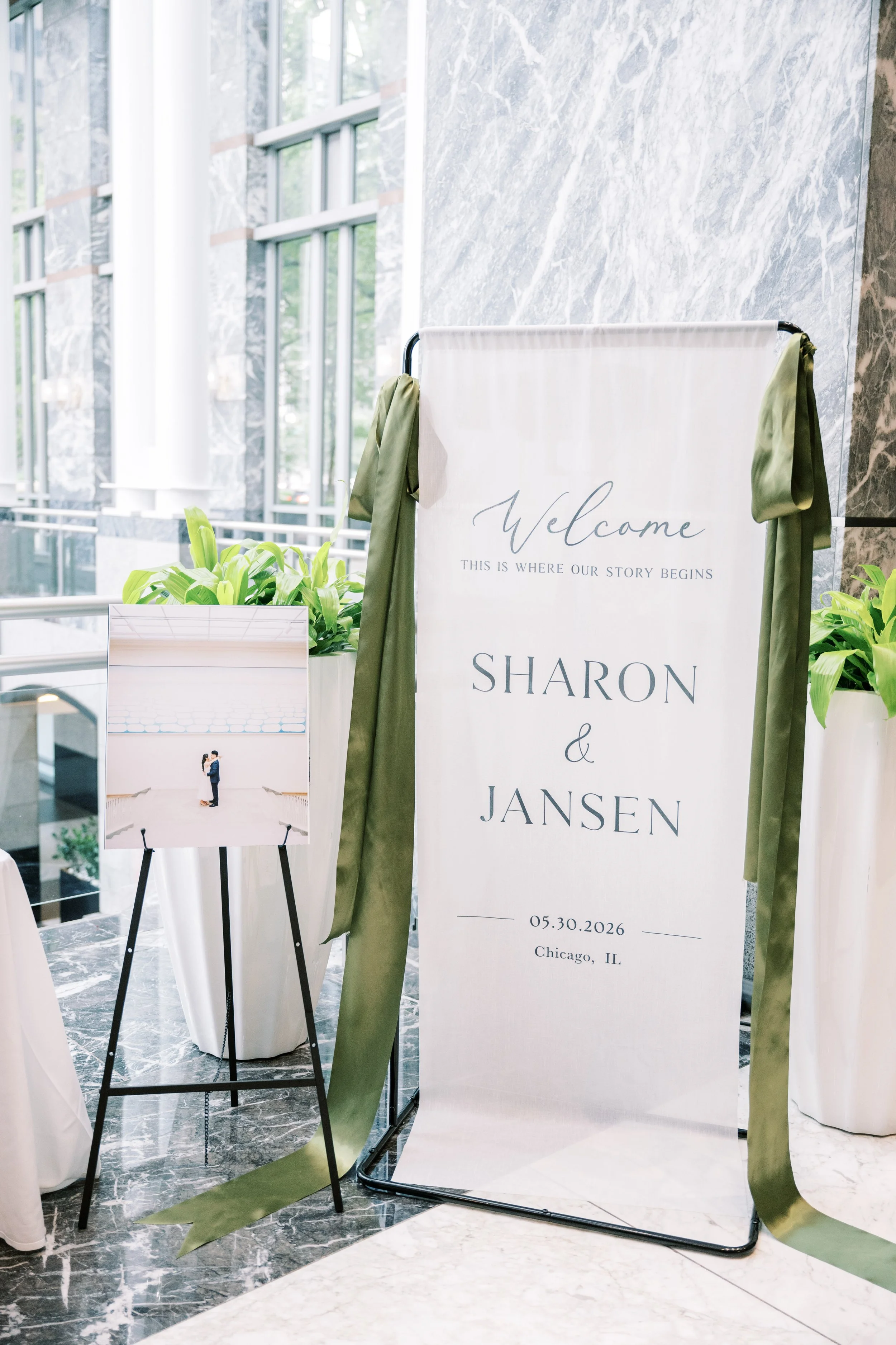 A large white wedding sign with green ribbons, displaying the names 'Sharon & Jansen', the date '05.30.2026', and location 'Chicago, IL'. A small table with a photo of a couple hugging and a green plant is nearby.