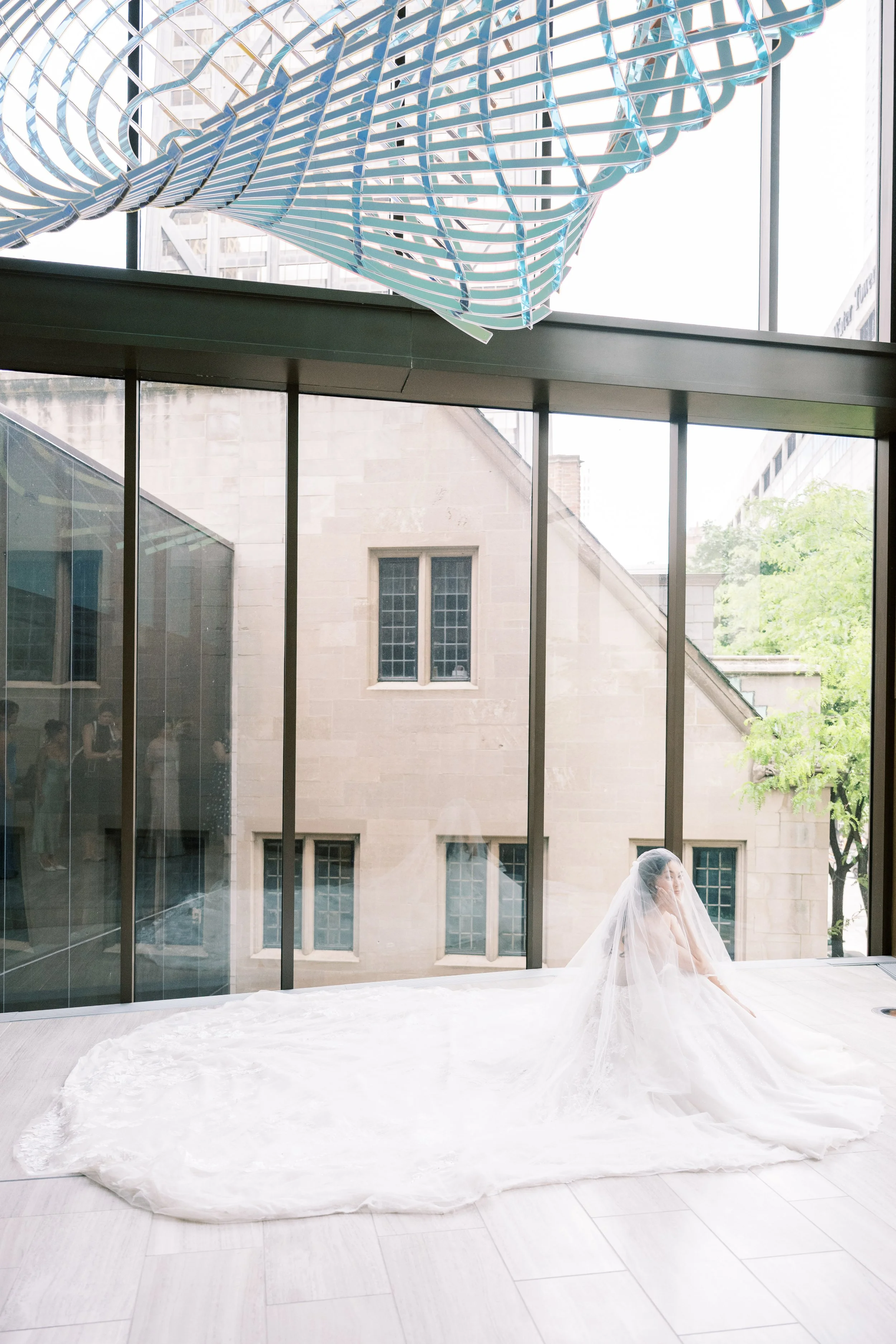 A bride sitting on the floor wearing a white wedding dress and veil, near a large window with a view of an urban cityscape with trees and old stone buildings.