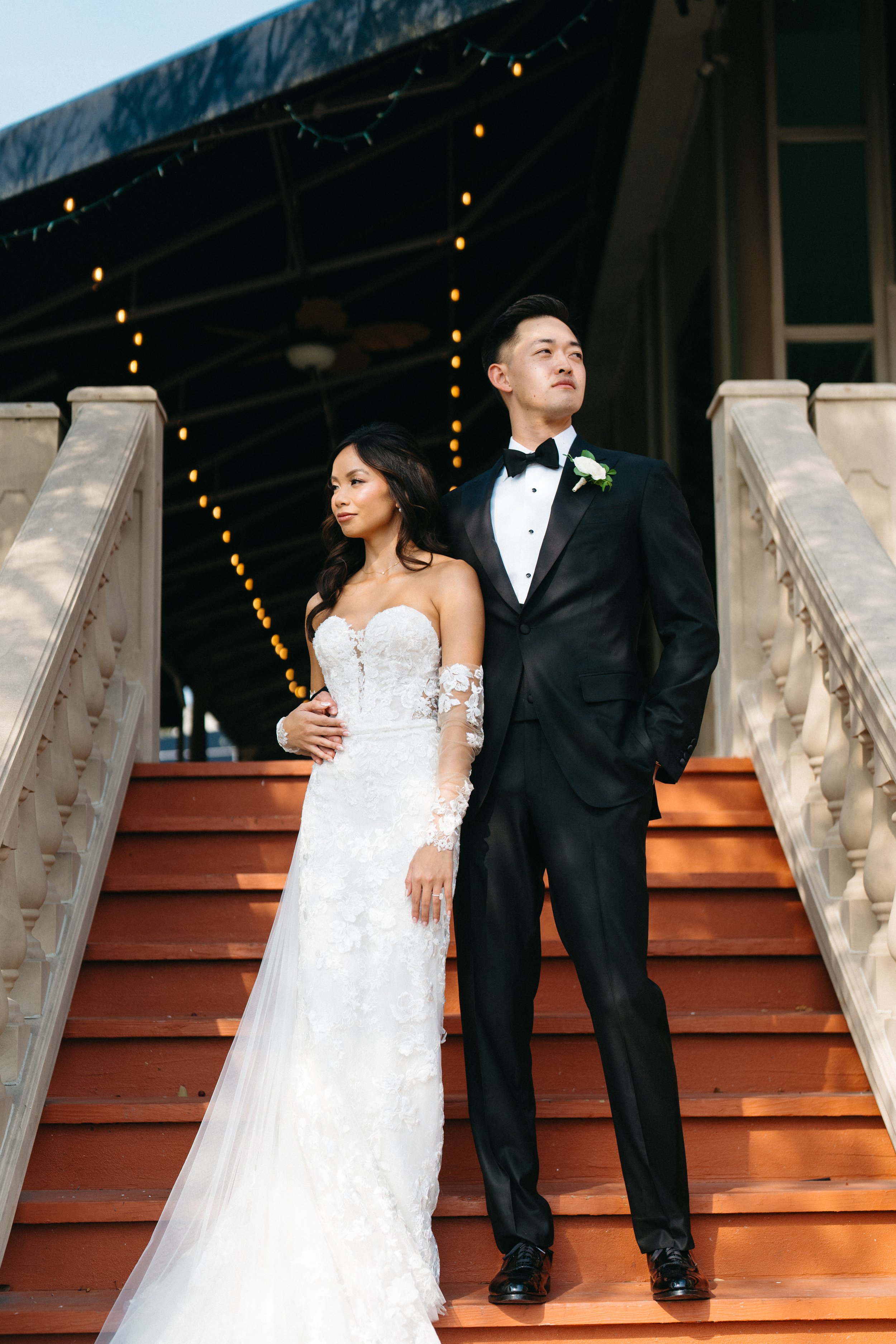 A bride and groom standing on wooden stairs outdoors during their wedding. The bride is wearing a white lace wedding dress with off-the-shoulder sleeves and a train, and the groom is wearing a classic black tuxedo with a bow tie and boutonniere. Ther