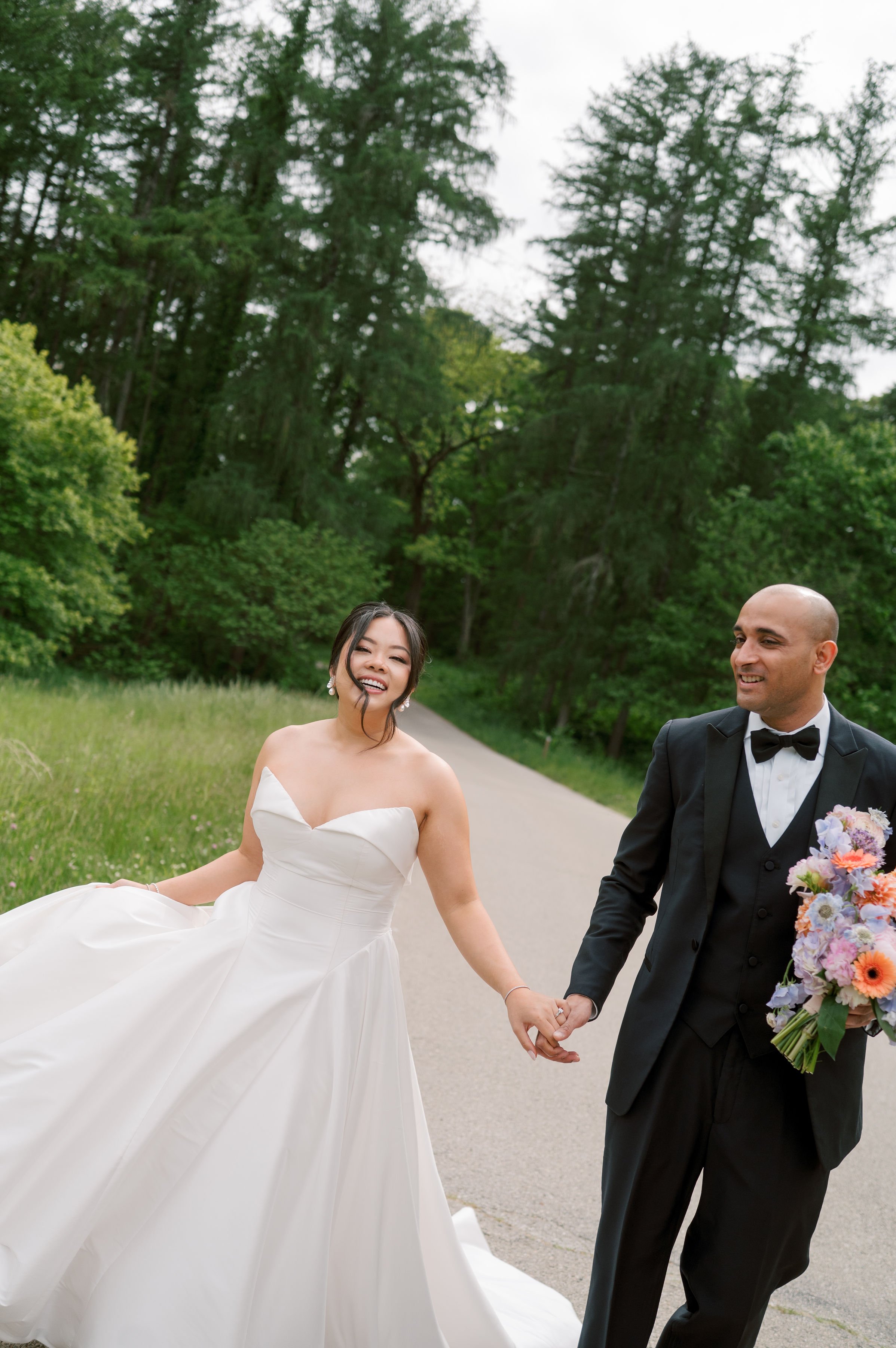 A bride and groom holding hands and walking outdoors on a paved path surrounded by green trees. The bride wears a white strapless wedding gown, and the groom is in a black tuxedo with a bow tie and boutonniere.
