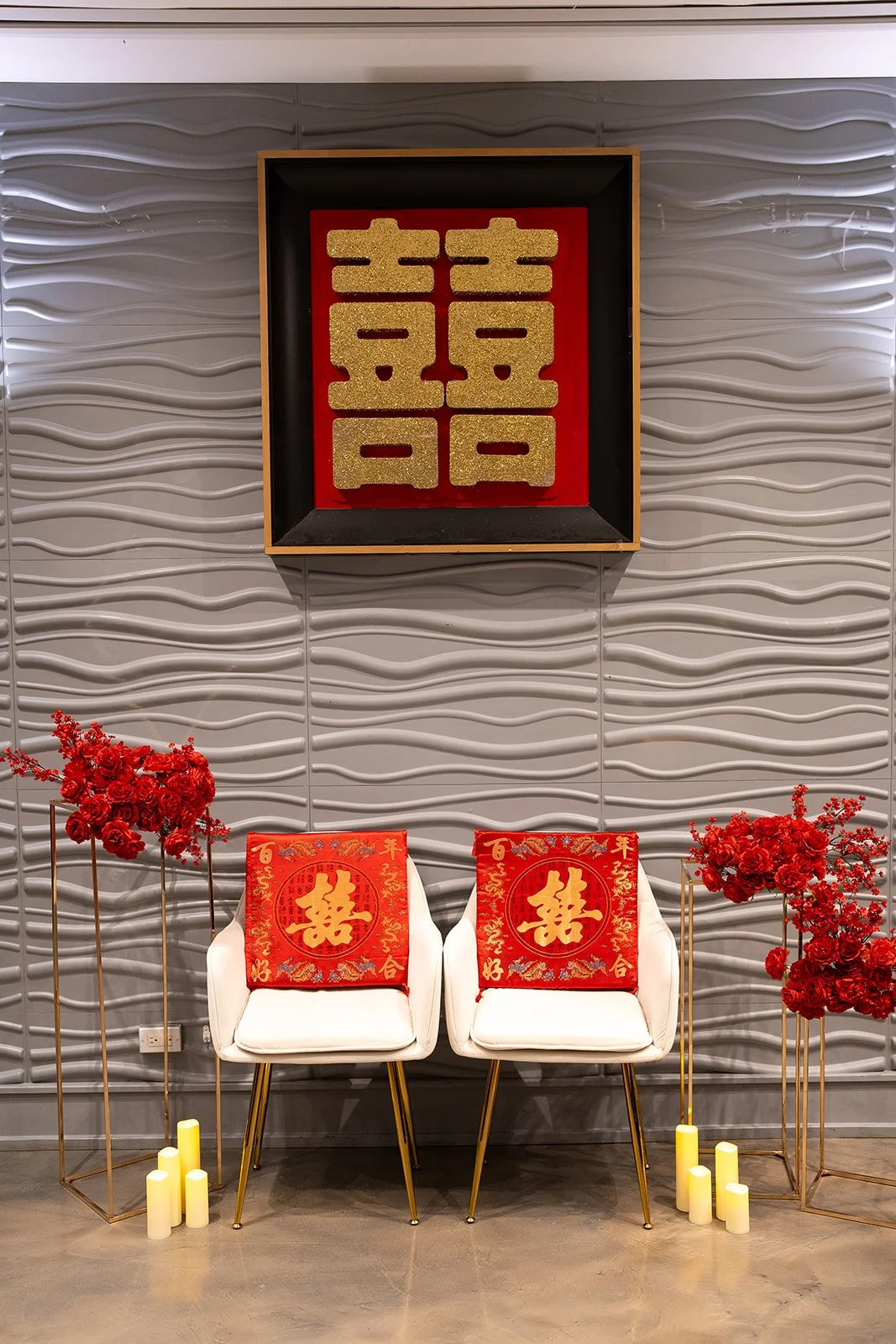 Decorations for a Chinese wedding or celebration featuring double happiness symbols on red banners, red flowers, candles, and a framed Chinese character for double happiness on the wall.