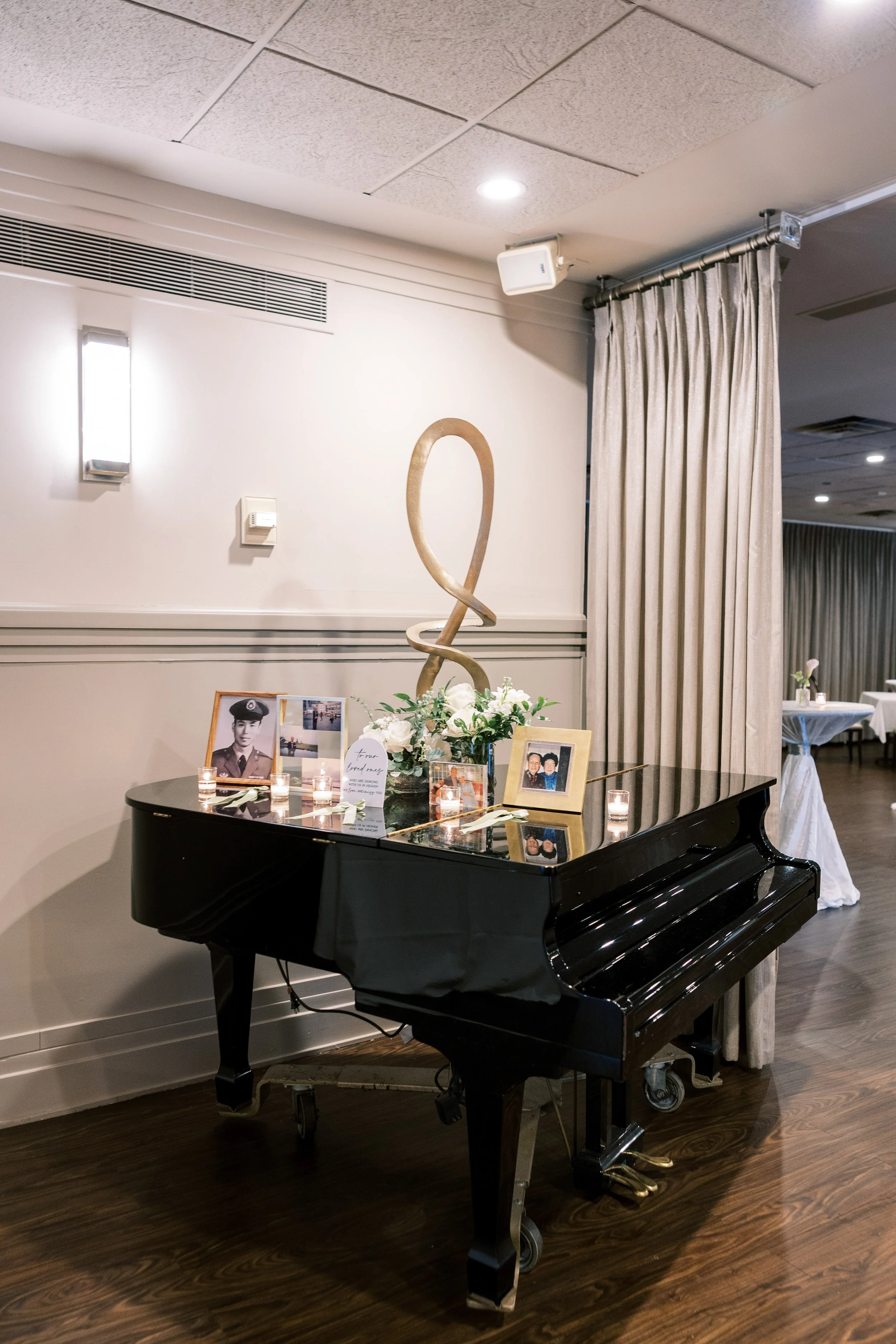 A black grand piano with framed photographs, candles, and floral arrangements on top, situated in a decorated event space with a white wall, beige curtain, and ceiling lights.