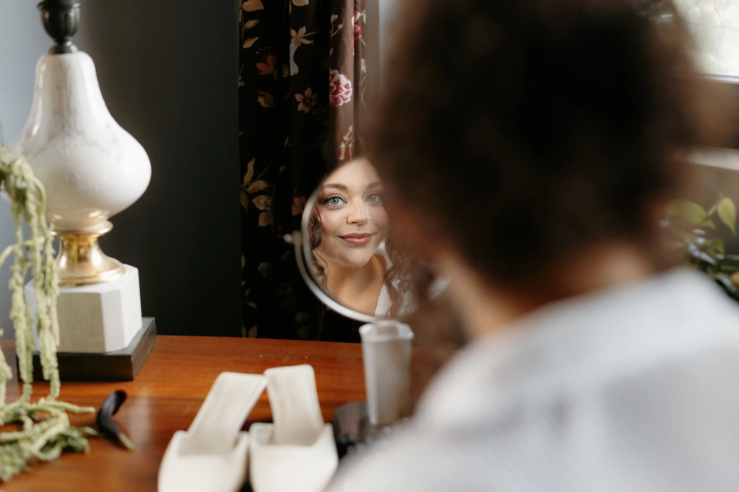 A woman is looking into a mirror, with her reflection clearly visible, while her face is partially obscured by a blurred object in the foreground. The setting appears to be a room with floral curtains and a table with various items, including a white