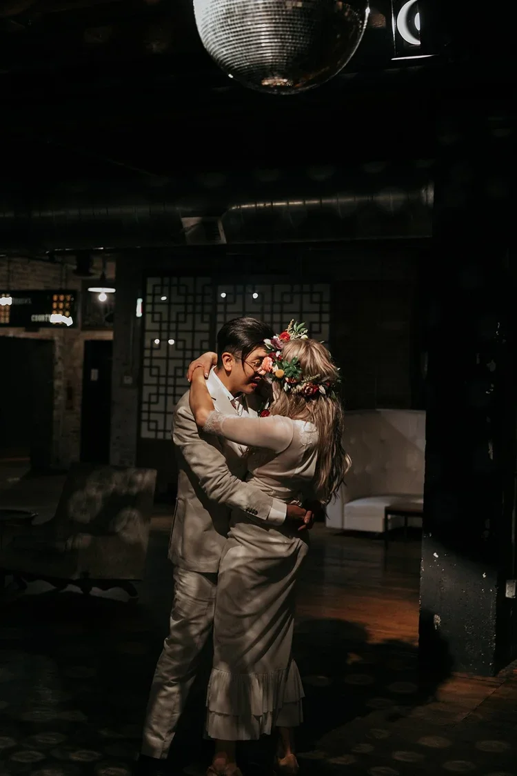 A couple dancing closely in a dimly lit room with a disco ball overhead, the woman wearing a flower crown and the man in a suit.