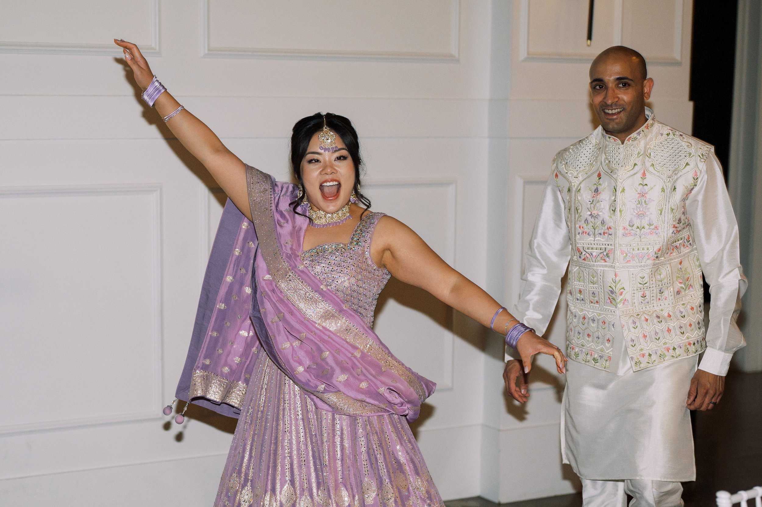 A woman in a traditional pastel purple and gold Indian outfit dancing joyfully, with a man beside her in white embroidered traditional Indian attire at an indoor event.