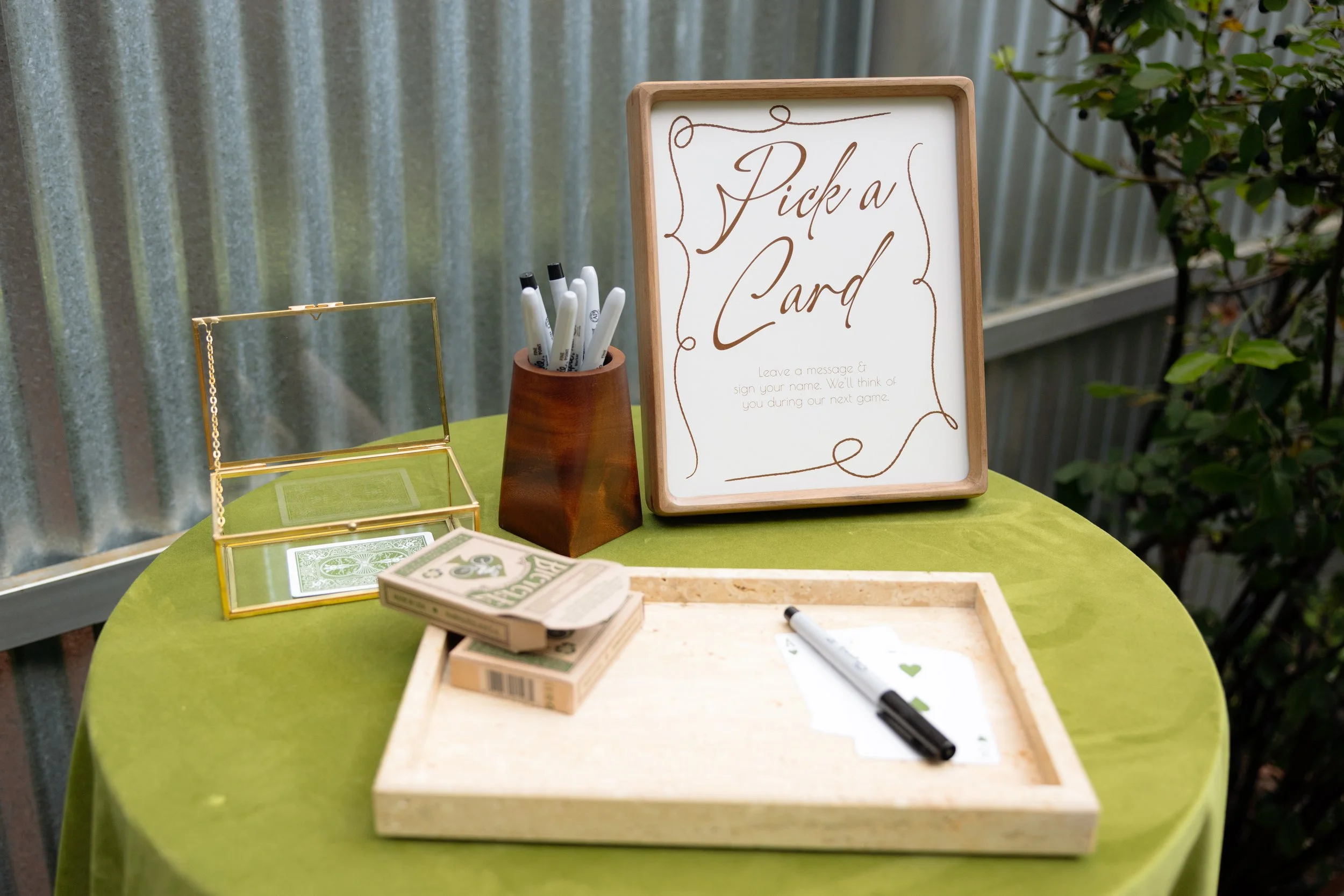 Sign reading 'Pick a Card' on a table with a deck of playing cards, a pen, and a small box, set outdoors with greenery in the background.
